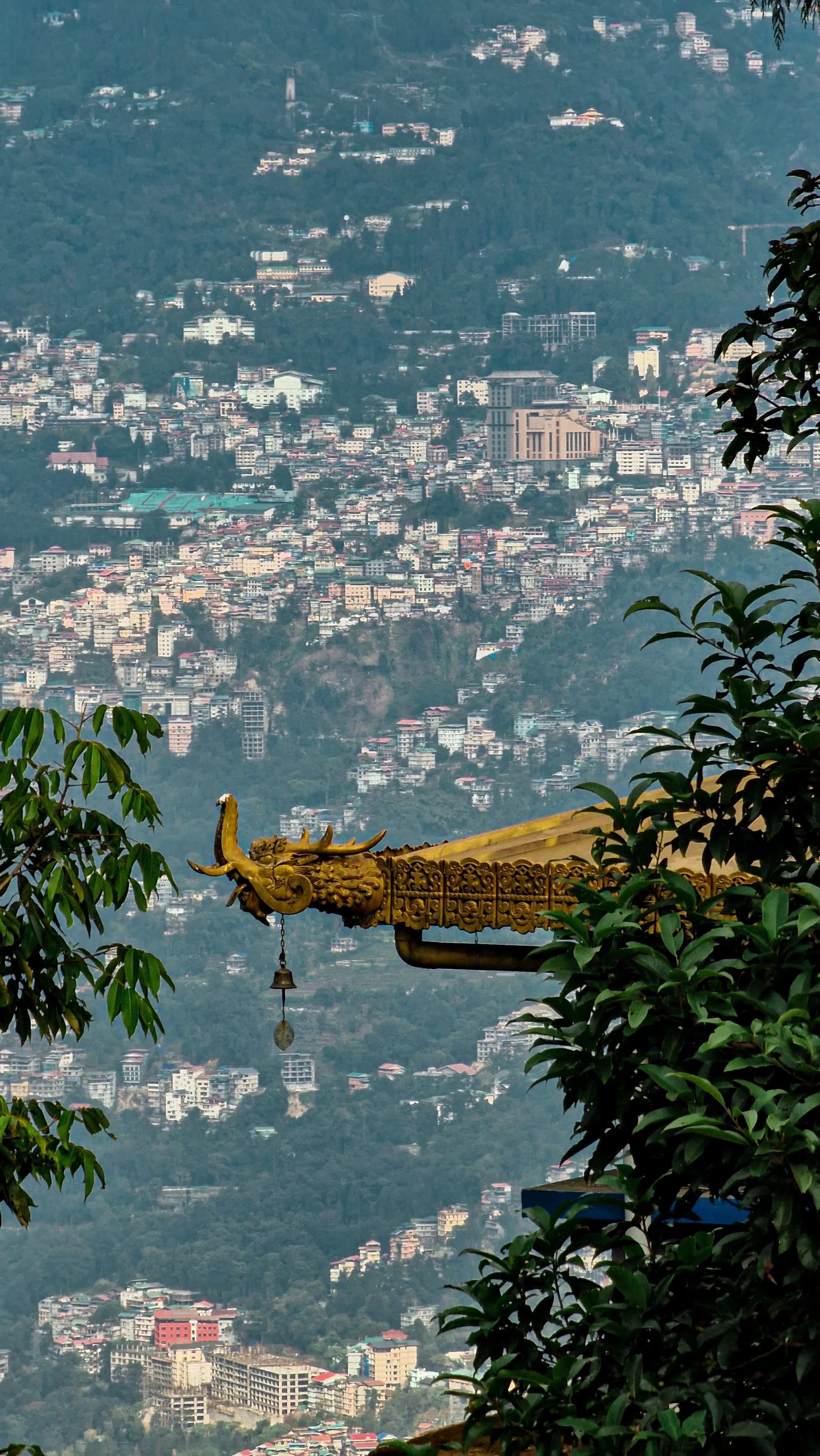 Cityscape view of a city in a hilly area with numerous buildings, partly obscured by green foliage. A decorative golden dragon sculpture with a bell is visible in the foreground.