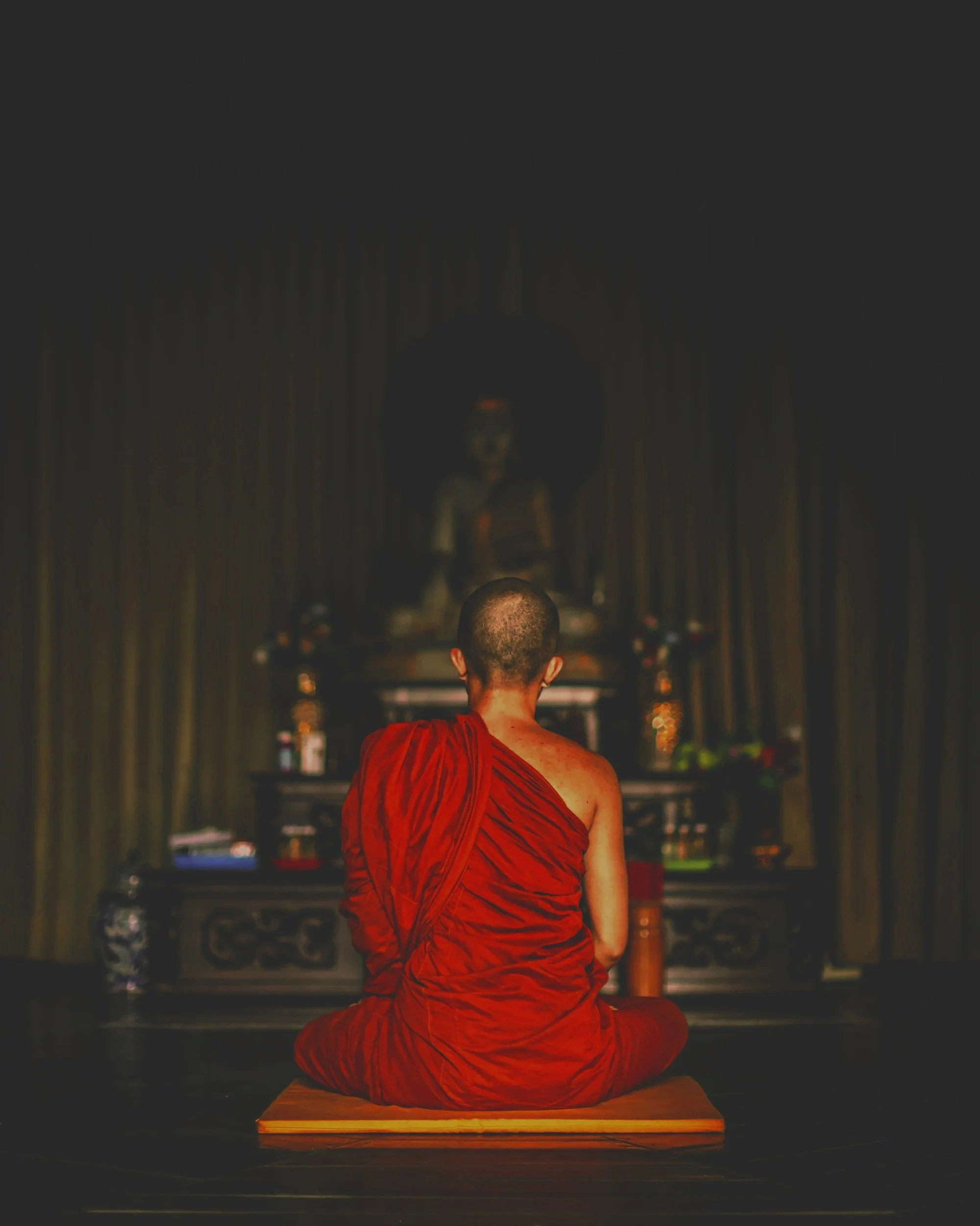 A Buddhist monk in a red robe sitting cross-legged on a wooden platform, facing a small altar with candles and spiritual objects, with a person in the background standing and observing.