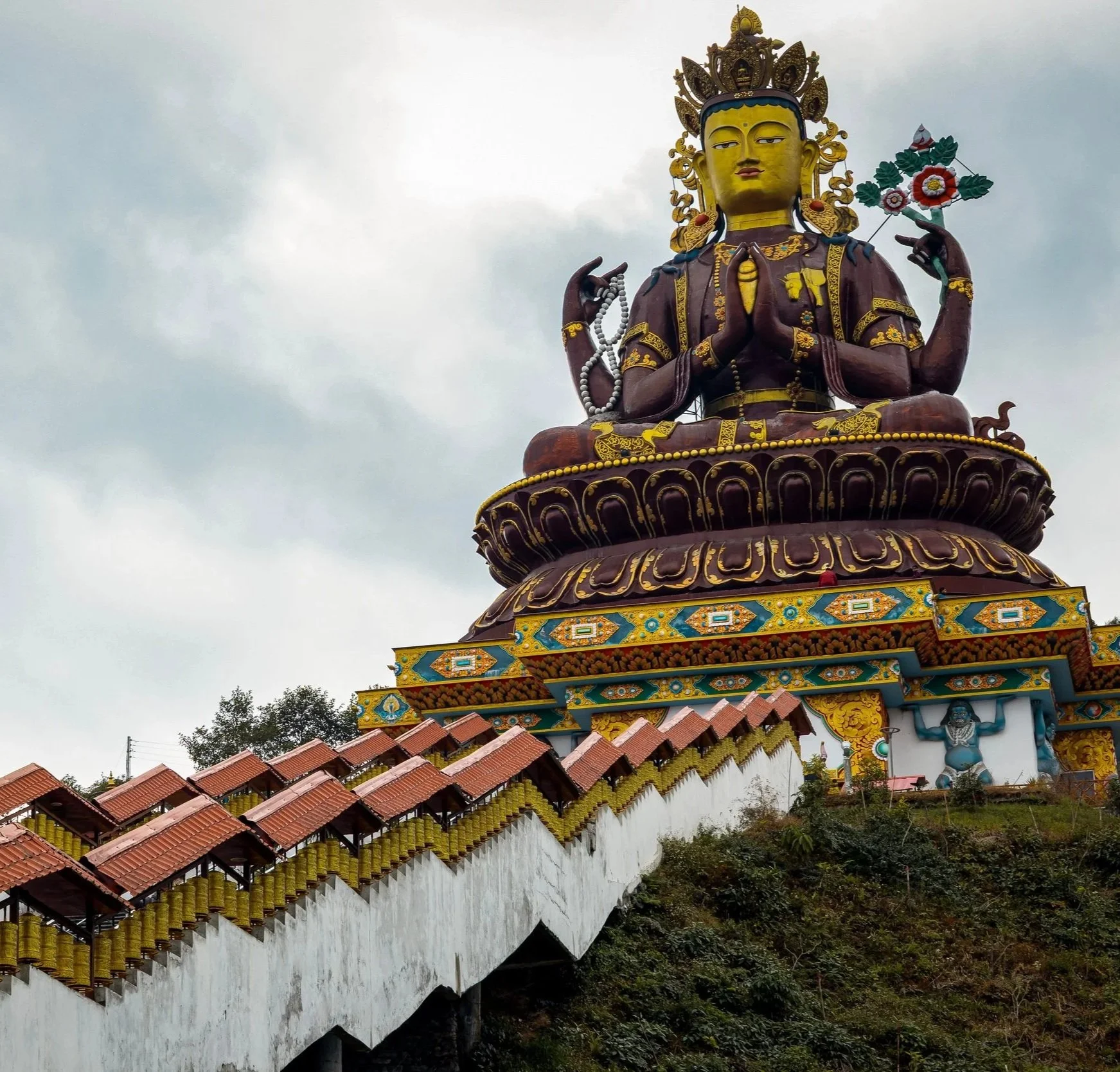 Huge colorful statue of a Hindu deity with multiple arms, seated on a decorated pedestal, with a backdrop of cloudy sky and palm trees, with a red-tiled roofed walkway in front.