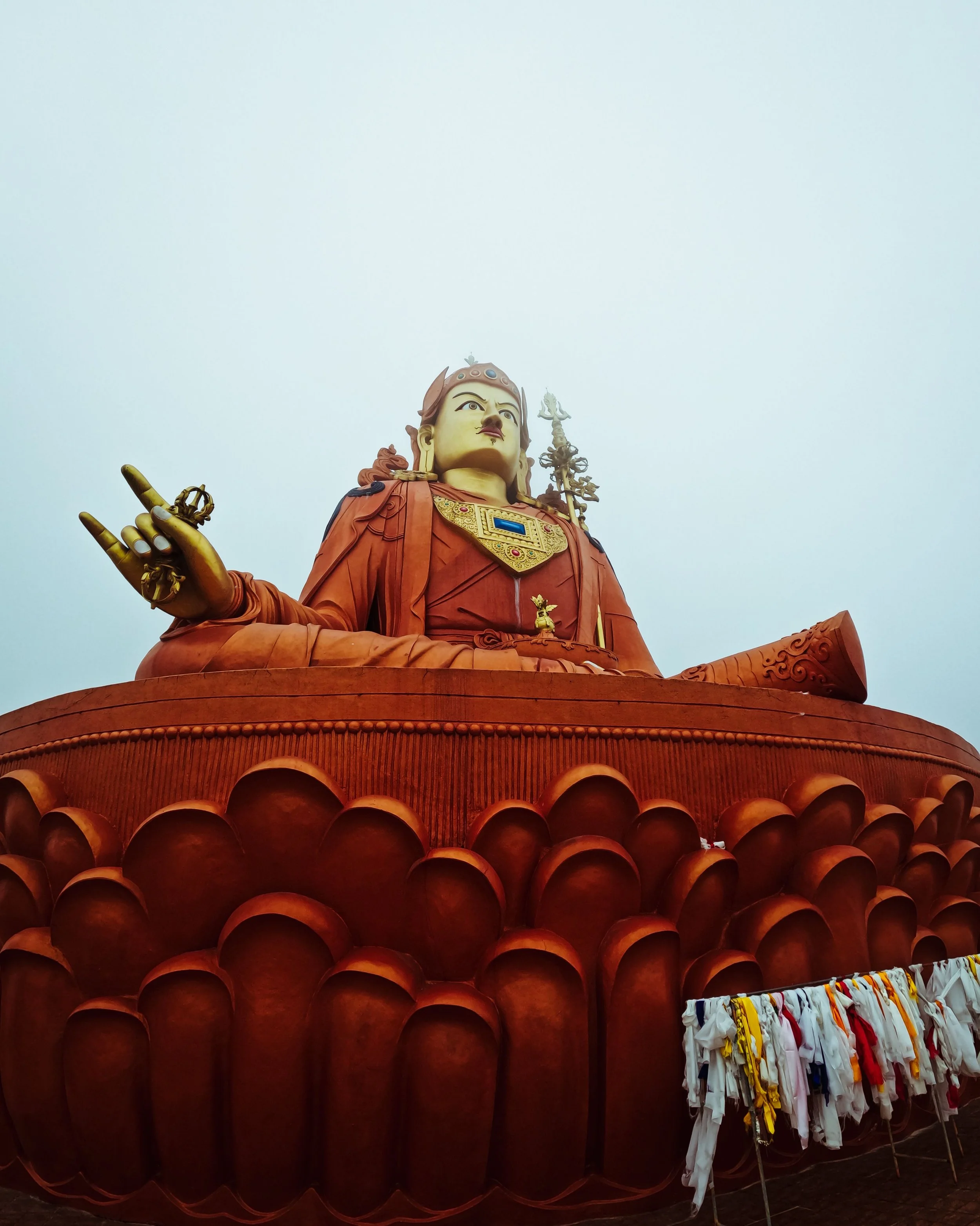 A large statue of a deity with multiple arms, adorned with elaborate jewelry and clothing, sitting on a lotus-shaped base. The statue is set against a cloudy sky.
