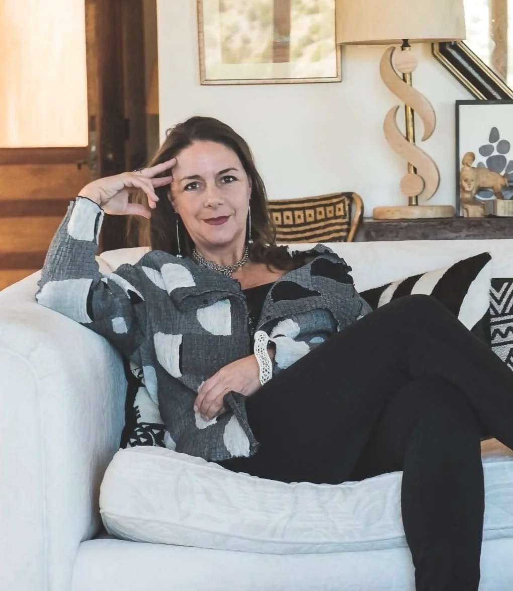 A woman with brown hair and a dark lipstick is sitting on a white couch, wearing a gray and white patterned jacket and black pants, with her right elbow resting on the armrest and her hand on her head, in a cozy room with artwork and a lamp behind her.