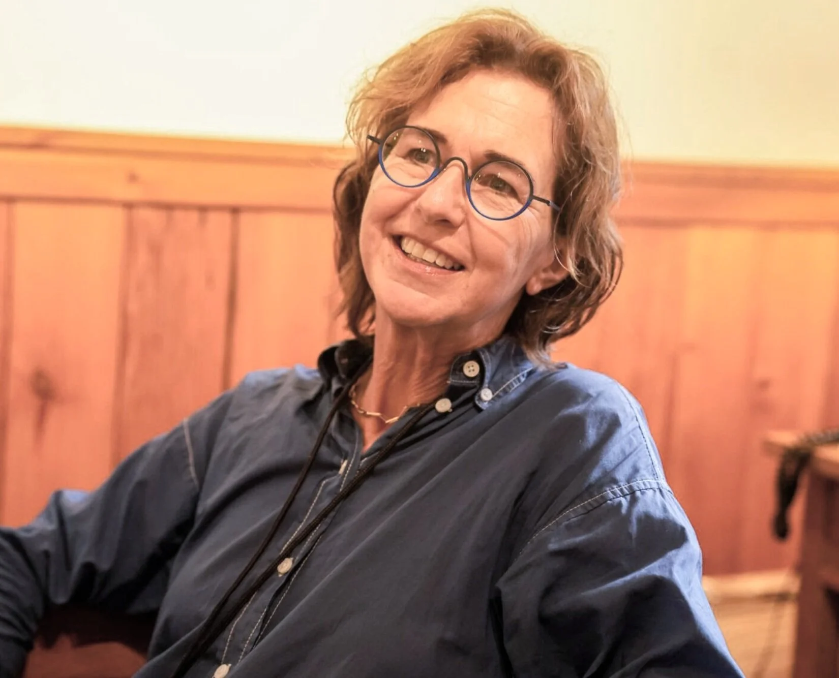 Woman with glasses smiling, seated indoors with wooden paneling on the wall behind her.