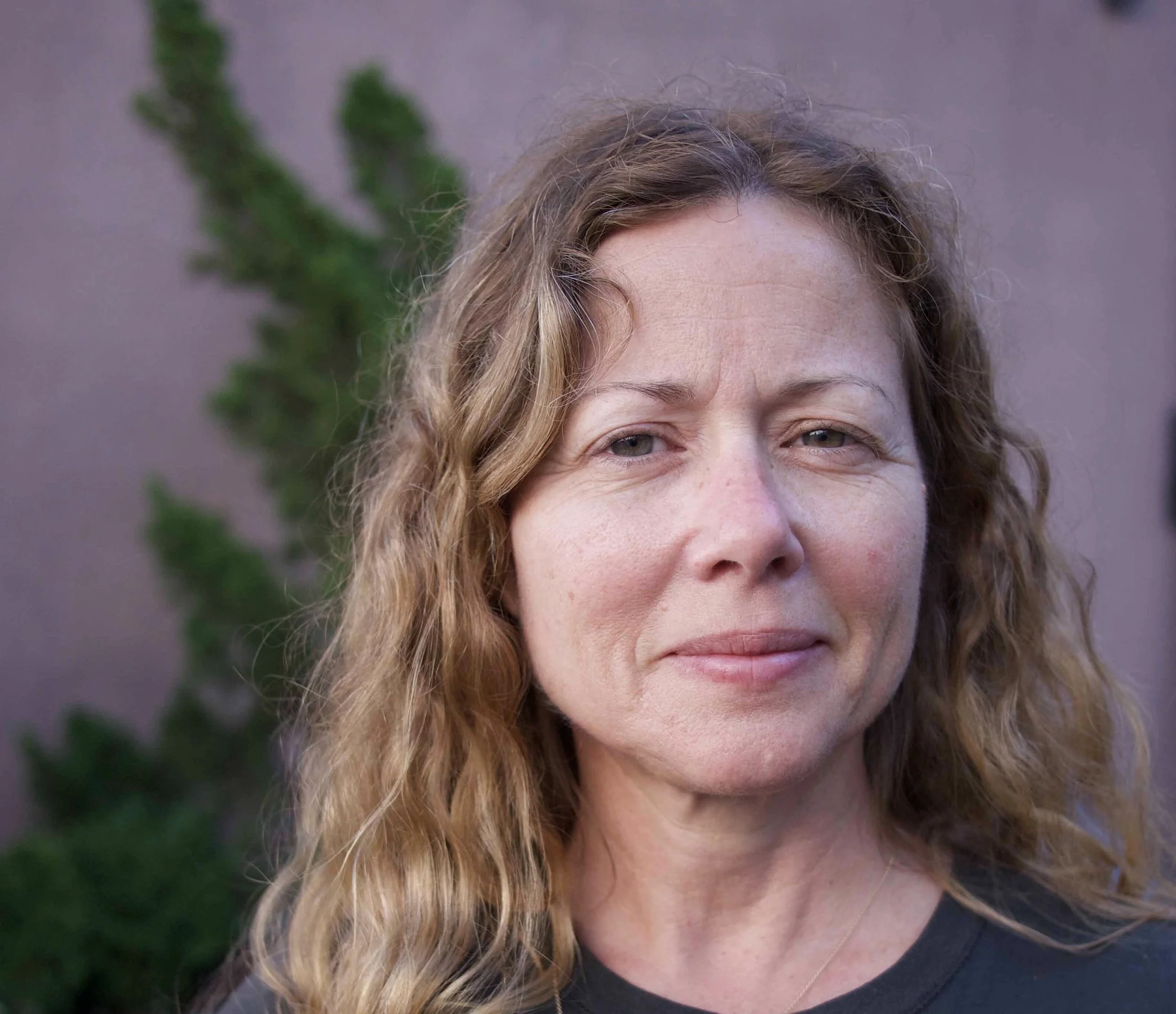 A woman with curly, reddish-brown hair and light skin, standing outdoors with a blurred green plant in the background.