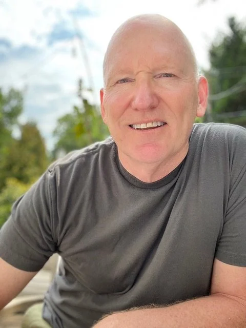 A smiling, bald man wearing a gray t-shirt outdoors with trees and power lines in the background.