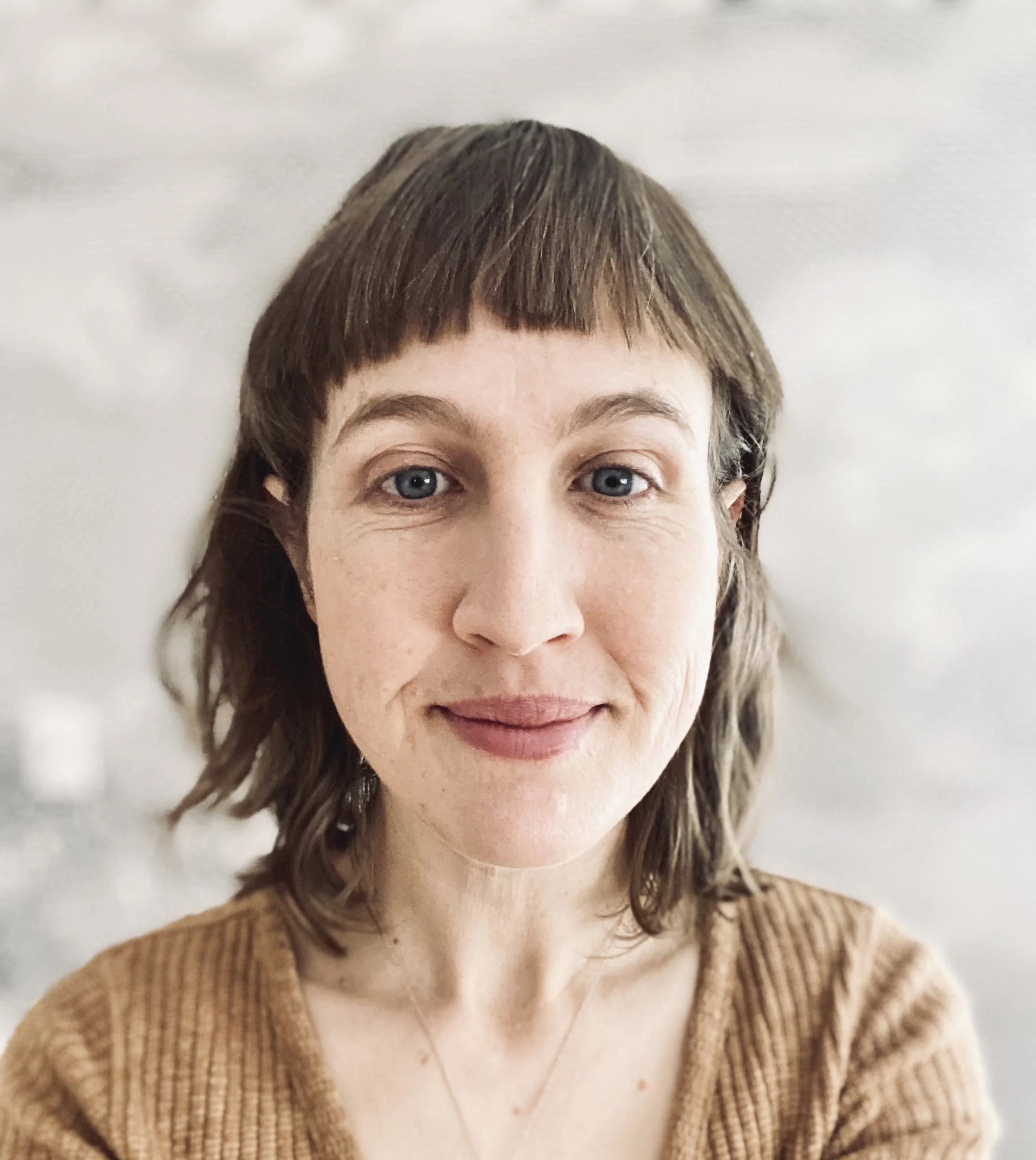 Close-up portrait of a woman with shoulder-length brown hair, blue eyes, and a gentle smile, wearing a brown sweater.