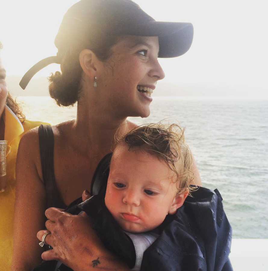 A woman with earrings and a baseball cap smiles as she holds a young child with wet hair, against a backdrop of water, likely on a boat.