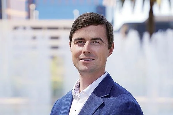 Portrait of a young man in a blue suit jacket and white shirt standing outdoors with a fountain and modern buildings in the background.