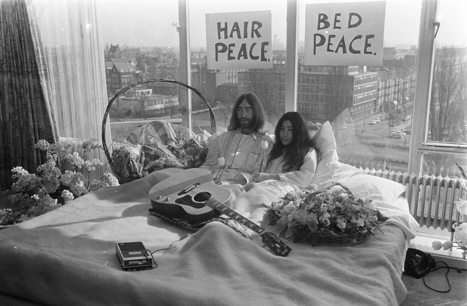 A couple lies in bed with flowers, a guitar, and a basket of flowers, in a room with large windows. Two signs in the window read 'Hair Peace' and 'Bed Peace.'