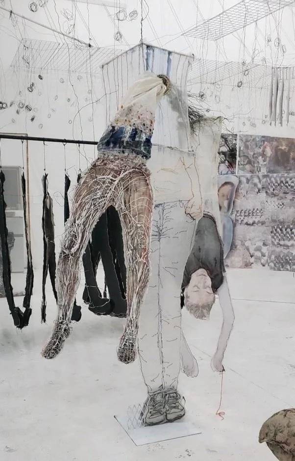 Mixed media art installation with suspended human and animal figures in a gallery space.