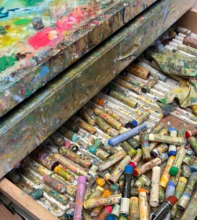 A messy artist's workspace with a stained paint palette on top and a drawer of used, paint-smeared oil pastel sticks scattered inside.