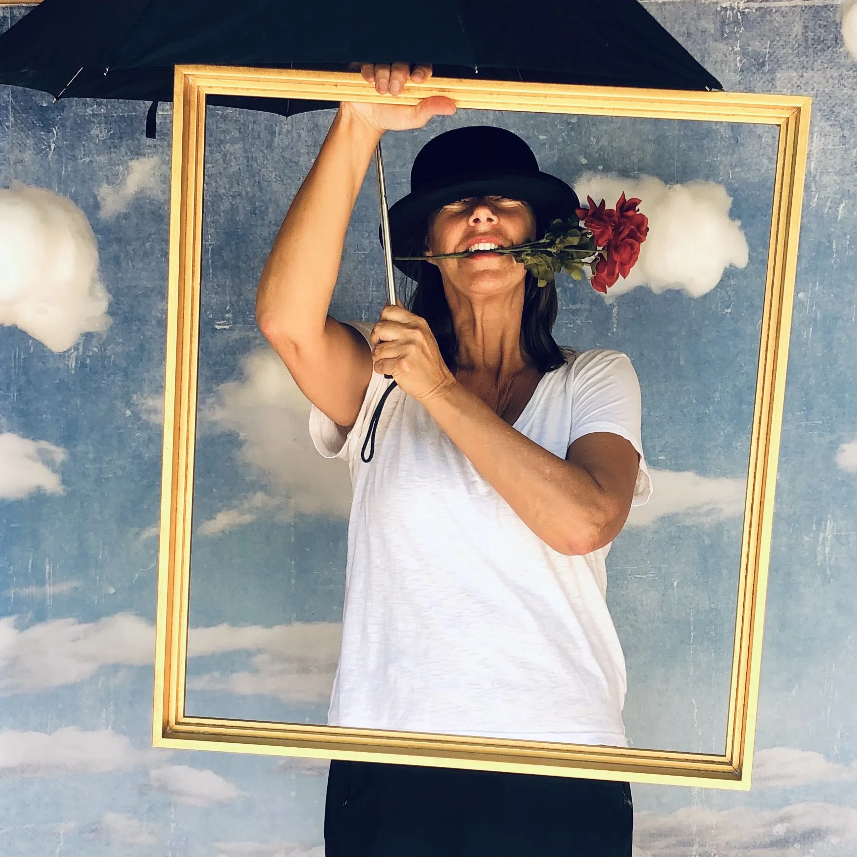 A woman wearing a black hat and white T-shirt holding a black umbrella, standing behind a large empty picture frame with clouds behind her, holding a red flower in her mouth.