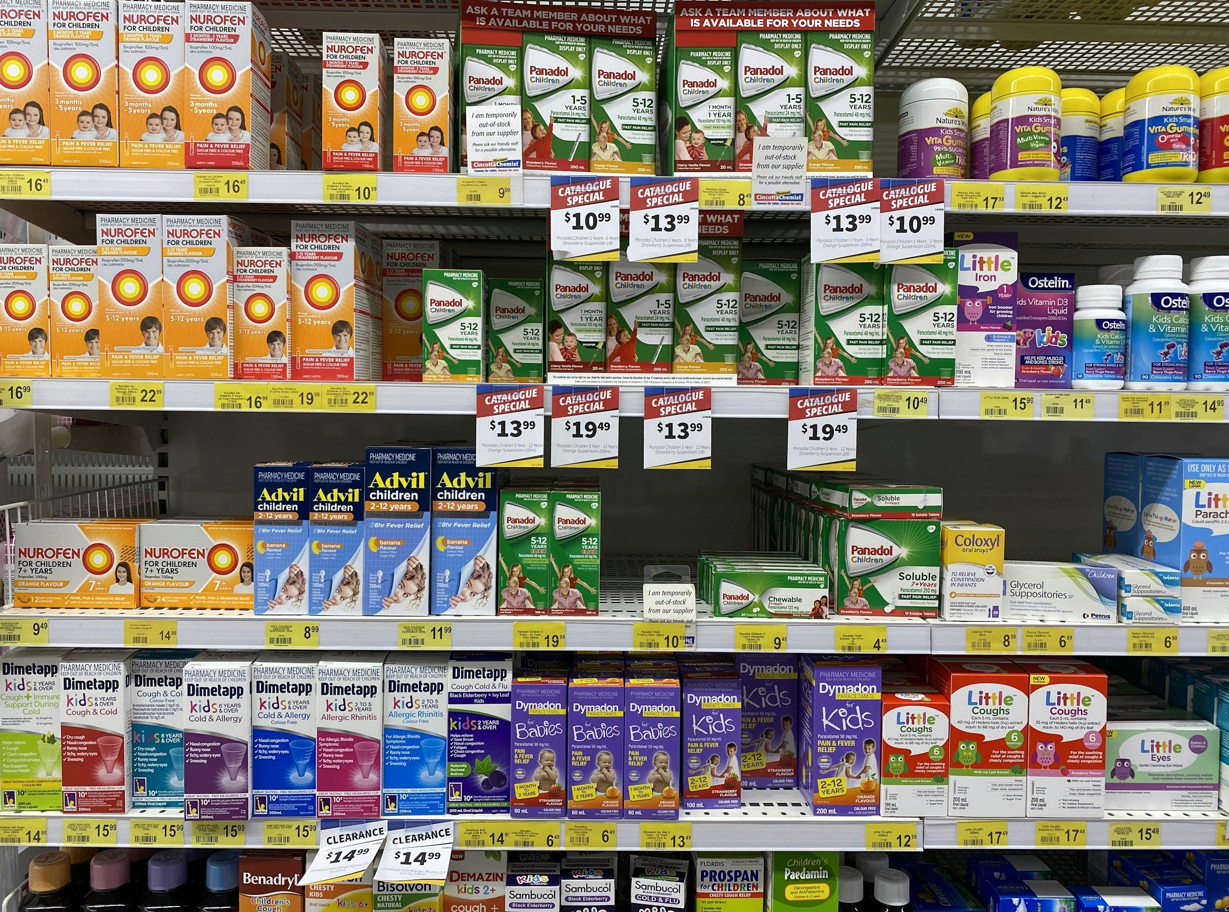 Shelf filled with various children's medication and vitamin boxes, including Nurofen, Panadol, Littlie Coughs, and Advil, with price tags and promotional signs.