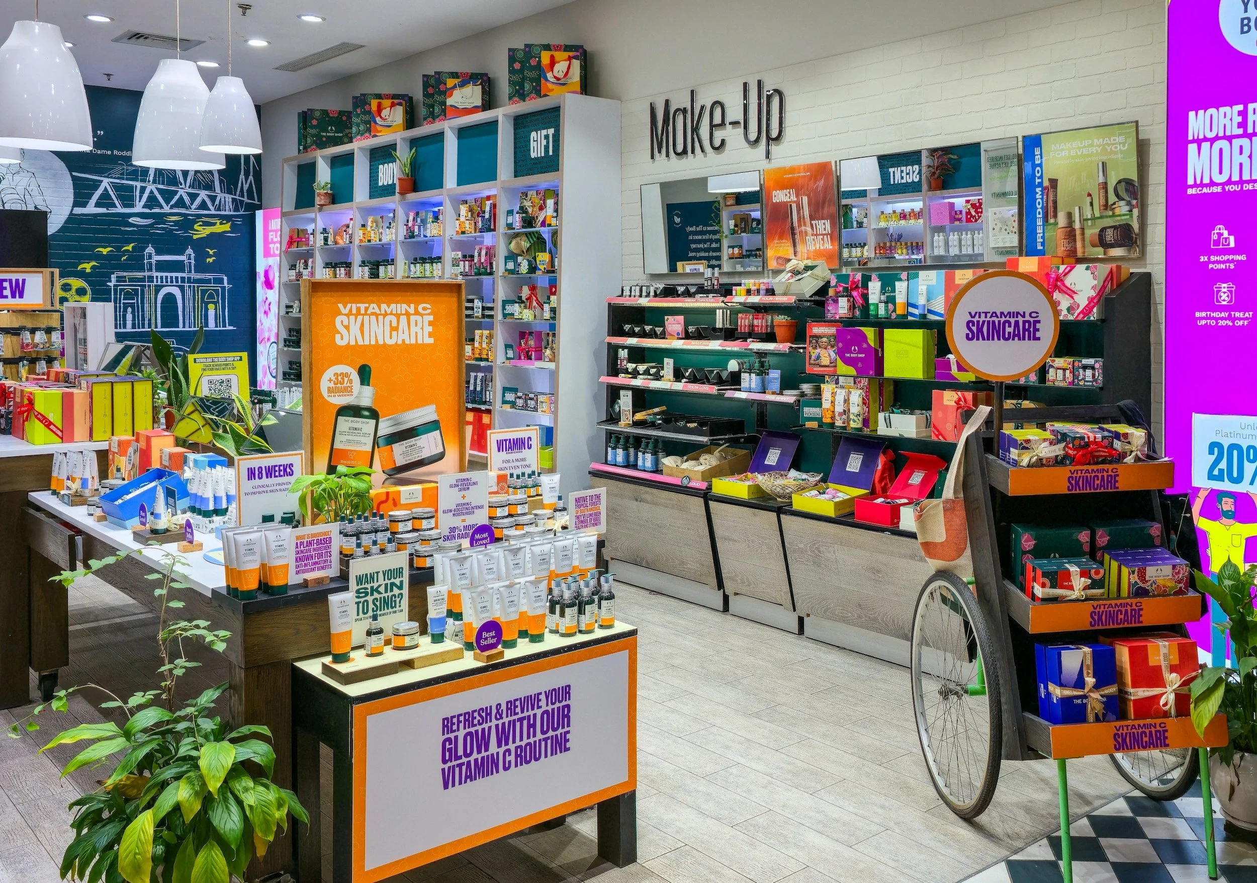 A retail store display for skincare products, featuring shelves with various bottles and boxes, including a sign promoting Vitamin C skincare routines, with a bicycle parked nearby and plants in the foreground.