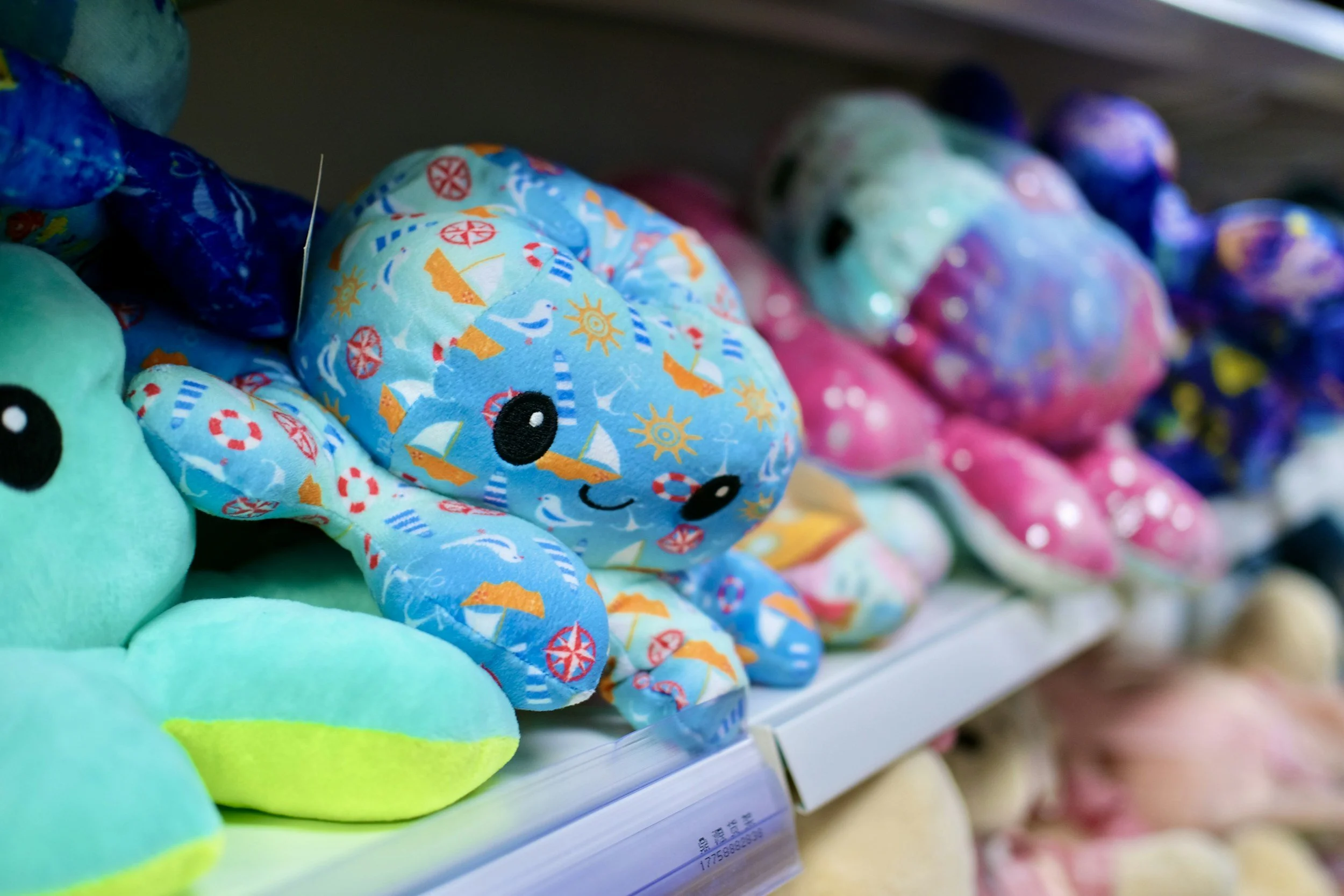 Colorful plush toys on store shelves, including a blue plush with sailboats and a smiling face, along with other soft toys in various colors.