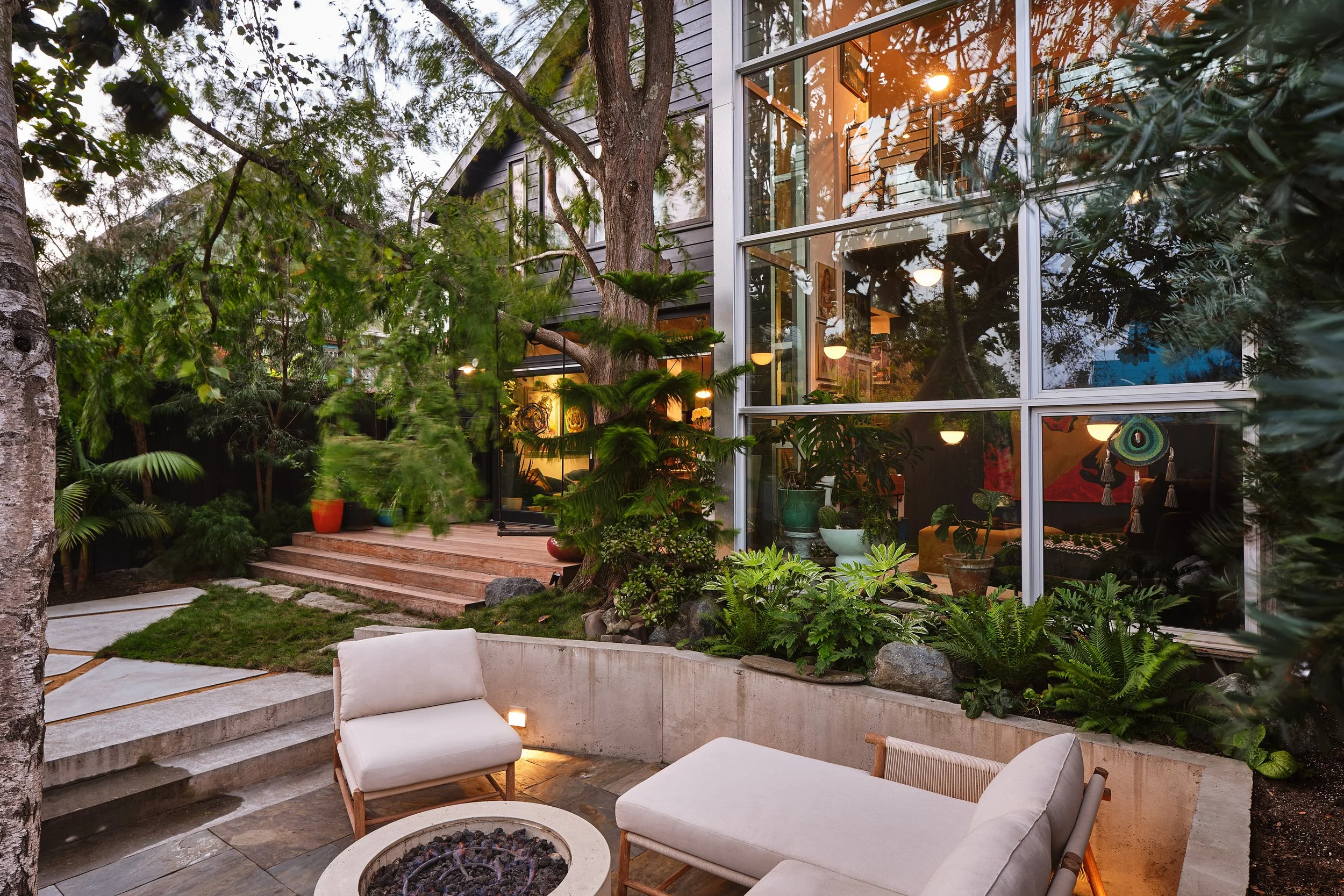 A modern backyard patio with outdoor furniture, lush greenery, trees, potted plants, and large glass windows reflecting trees and sunset