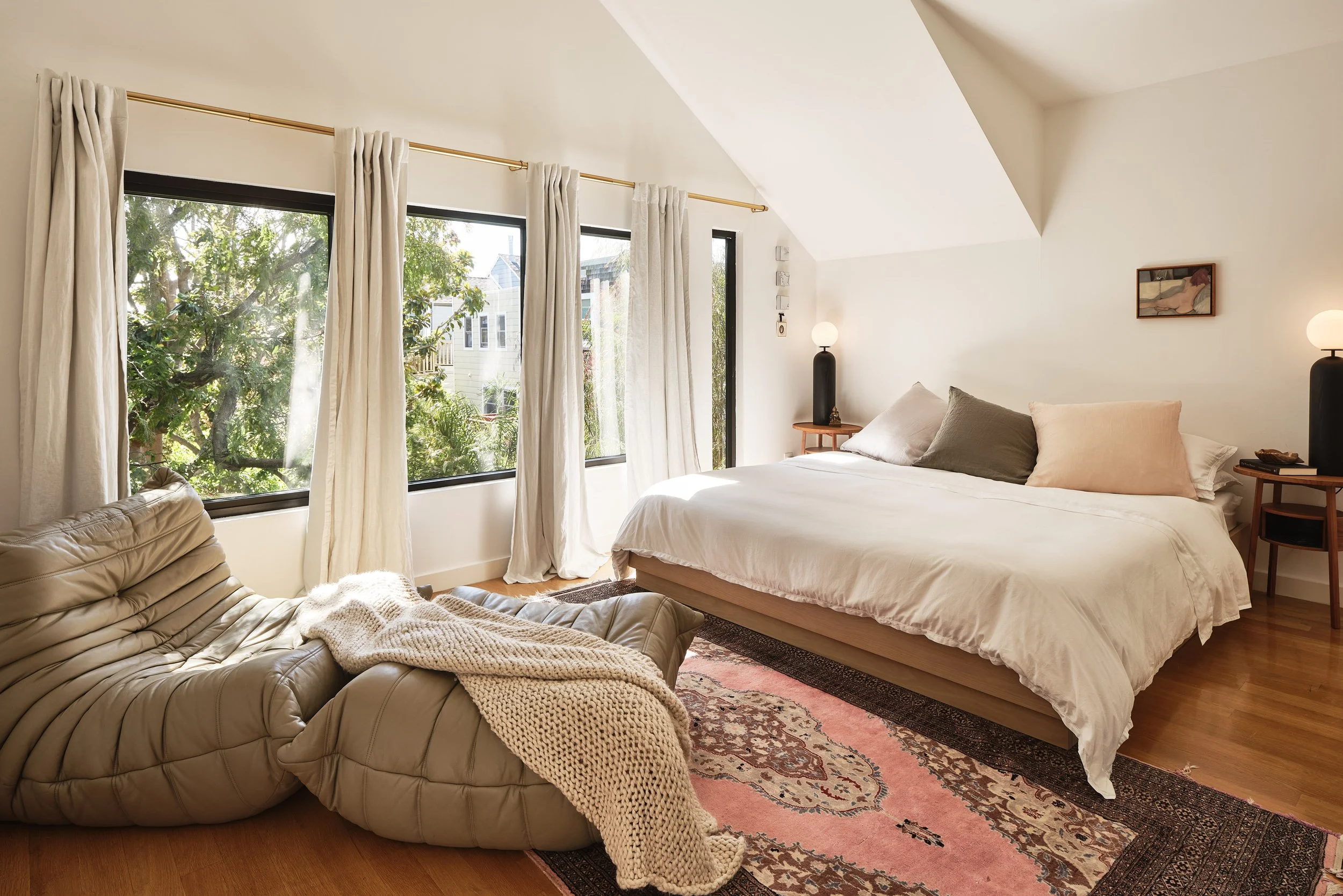 A bedroom with white walls and sloped ceiling, large windows with cream curtains showing trees outside, a bed with beige and gray pillows, a wooden nightstand with a lamp, and a wall picture of a nude woman. There is a cream leather sofa with a blank