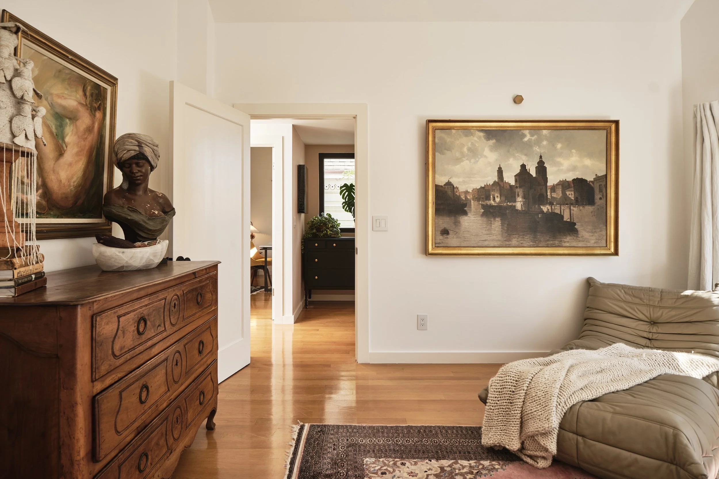 Living room with a wooden dresser, a bust sculpture, a large painting of a woman, a framed landscape painting, and a beige sofa with a blanket. A door opens to another room with a dark chest and a plant, and a window with curtains is on the right.