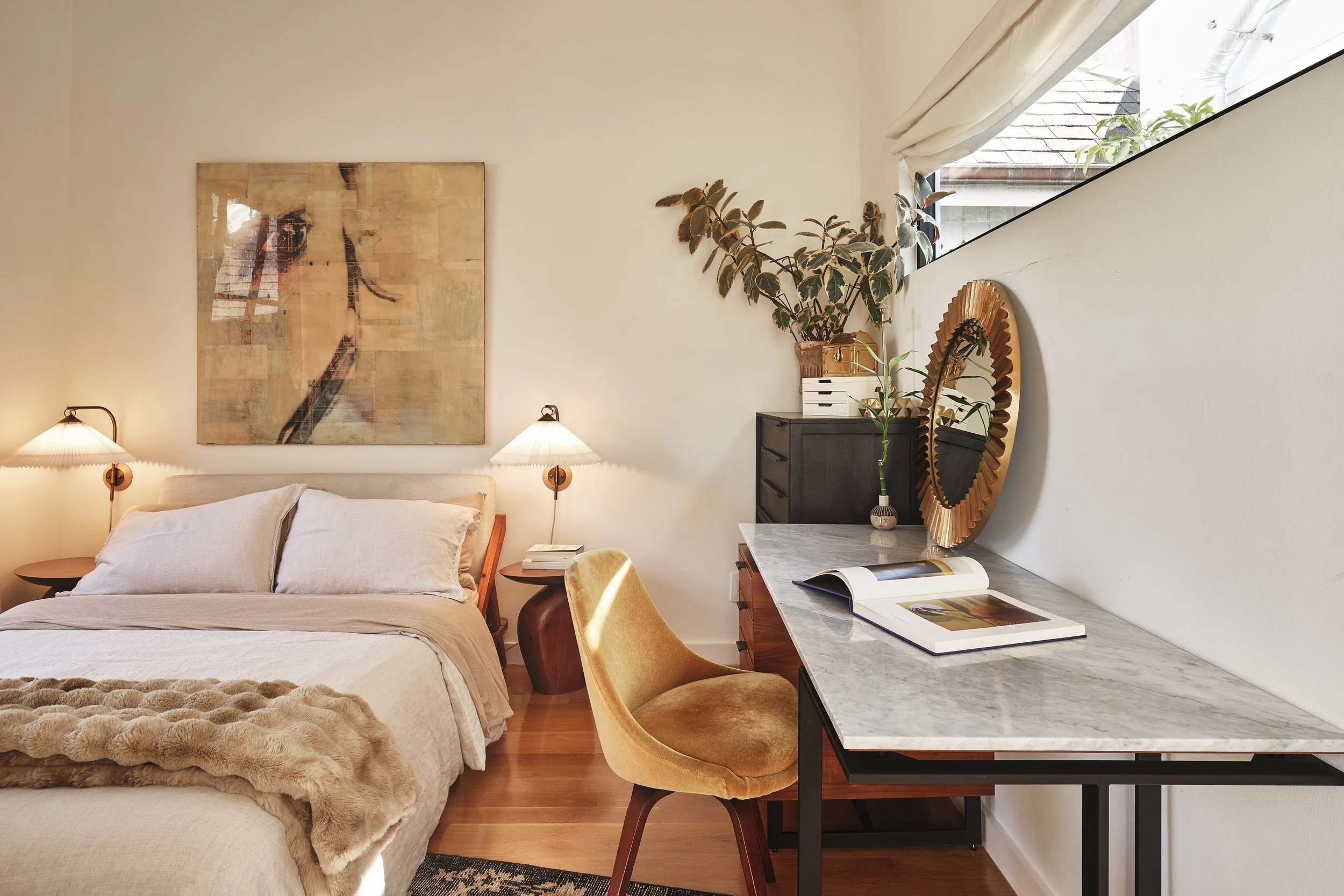 A cozy bedroom with a bed, two lamps, a large abstract painting, a dresser with decorative items, a mirror, and a marble-topped desk with a chair.