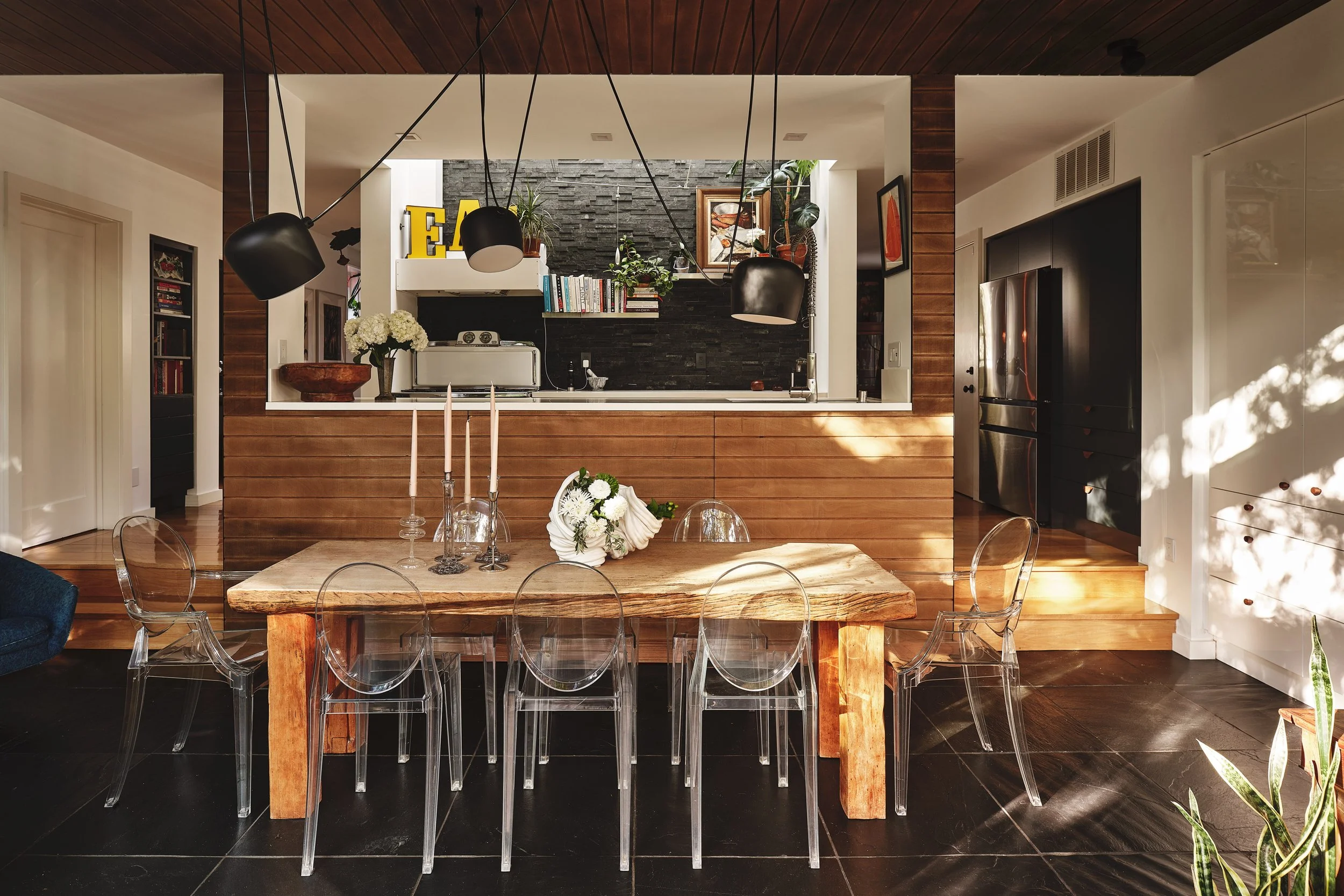 Modern dining room with a rustic wooden table, clear acrylic chairs, and black pendant lights. Visible in the background is an open kitchen with a black brick backsplash, shelves with books, plants, and decorative items. Sunlight is streaming into th