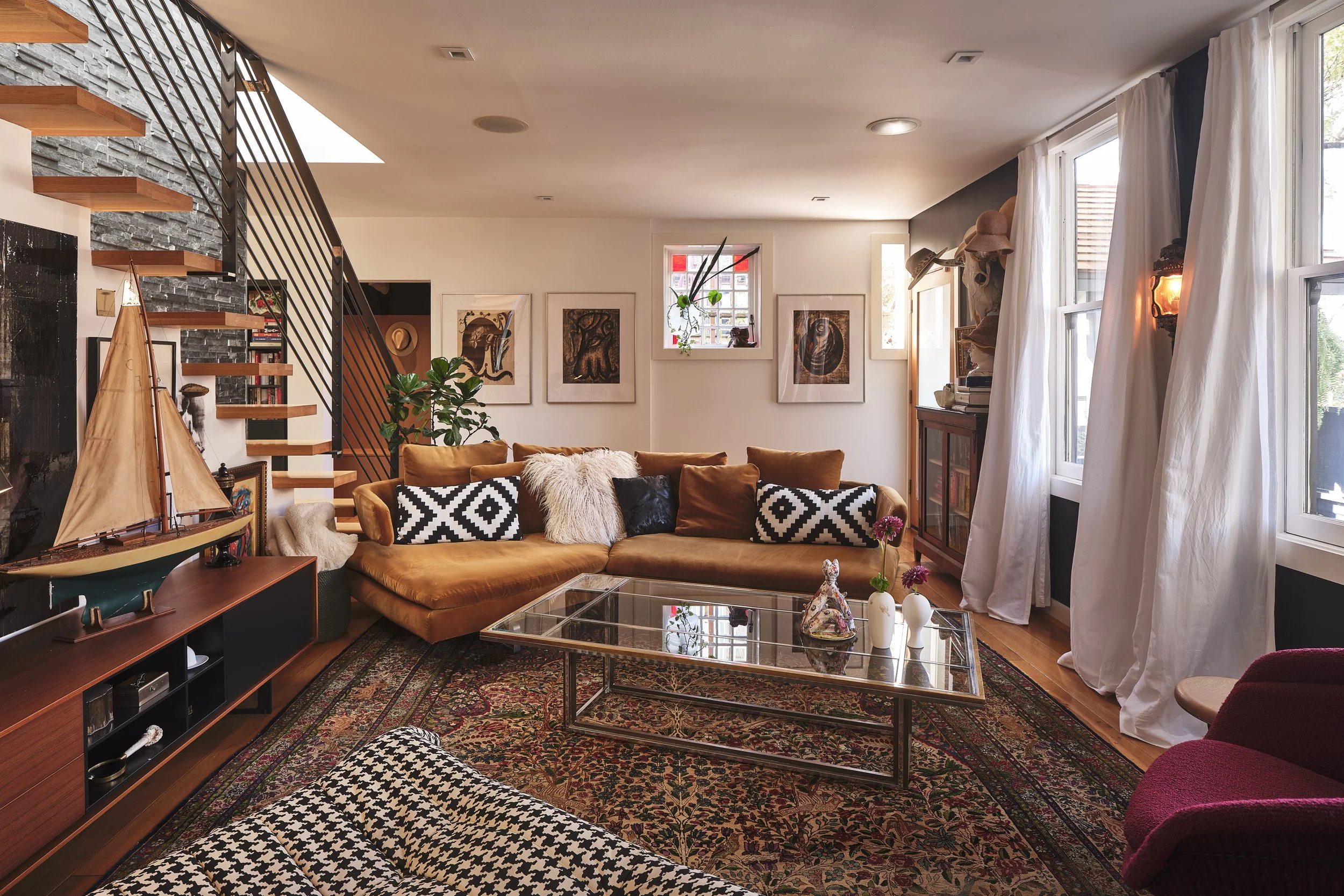 Living room with orange velvet sofa, black and white patterned pillows, glass coffee table, patterned rug, artwork on white walls, large windows with white curtains, various hats on a shelf, and indoor plants.