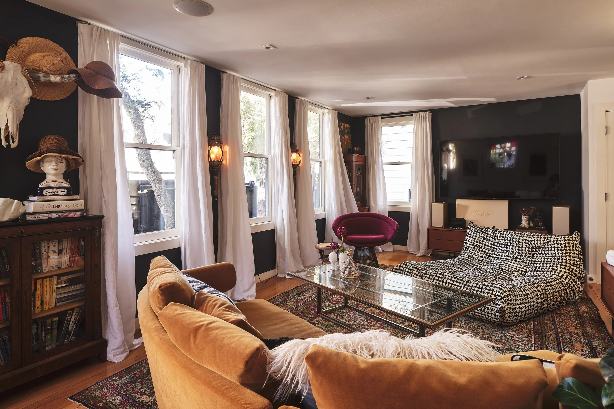 A cozy living room with multiple large windows draped with white curtains, black walls, and hardwood floors. The room features a beige velvet sofa, a black and white patterned armchair, a glass coffee table, and a vintage wooden cabinet. Decor includes colorful paintings, lamps, hats on a mannequin bust, and several books, creating an eclectic and inviting atmosphere.