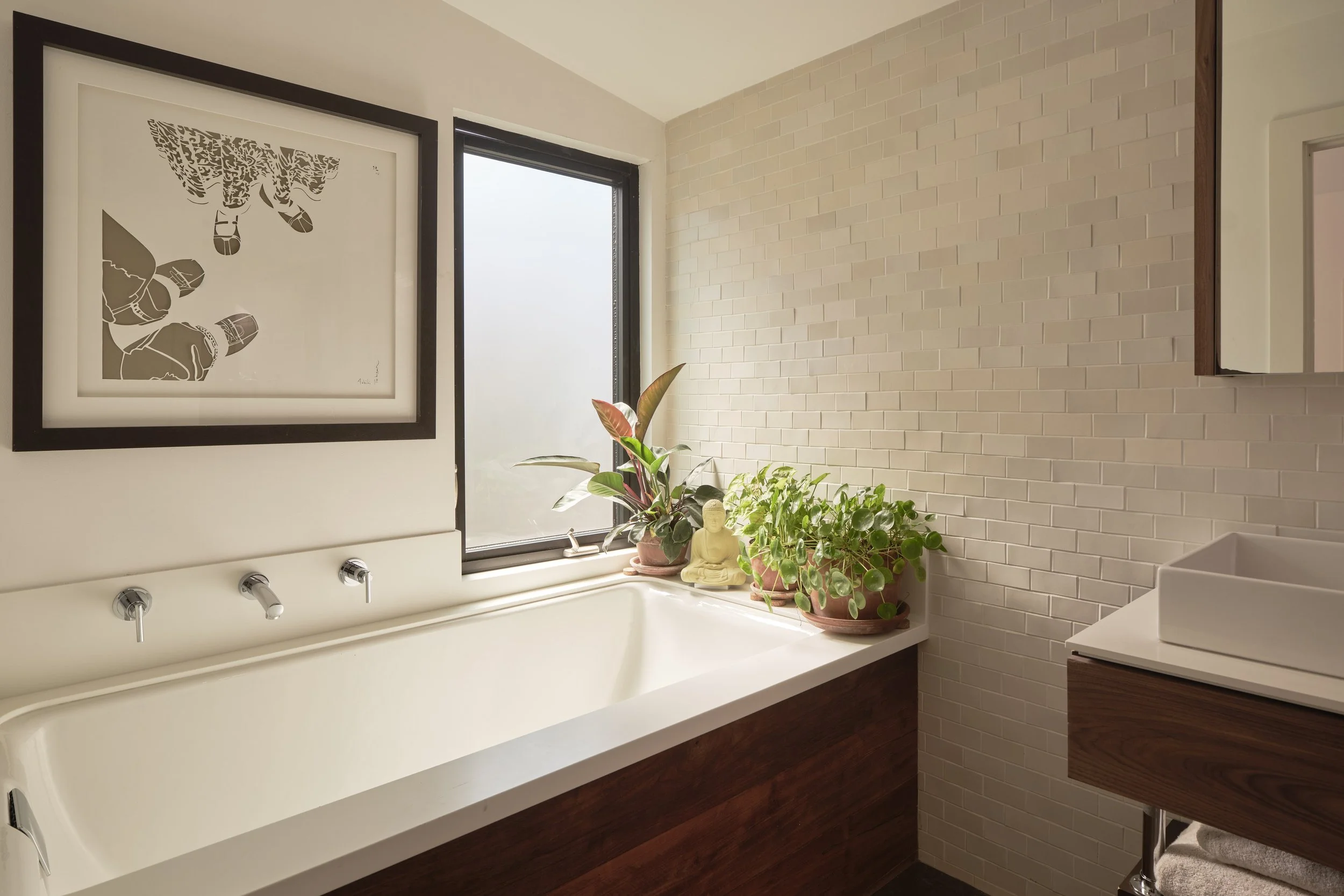 A modern bathroom with a white bathtub, potted plants on the edge, a Buddha statue, framed art of hanging shoes on the wall, and a textured white brick wall with a window.