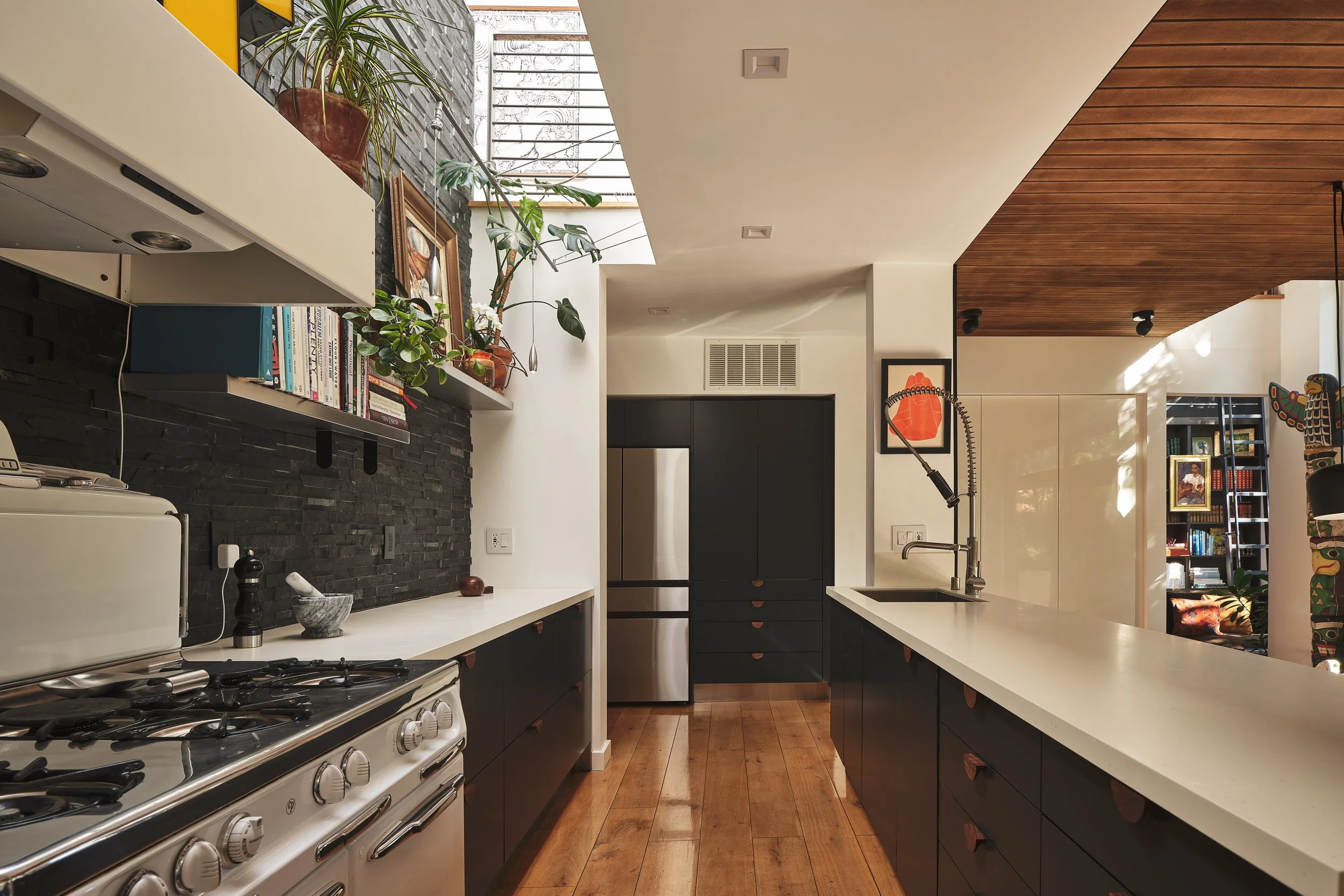 Modern kitchen with black and white cabinetry, a white countertop, and a stove. There are plants on a floating shelf and artwork on the wall. Natural light enters through large windows, with wooden floors and a wooden ceiling section.
