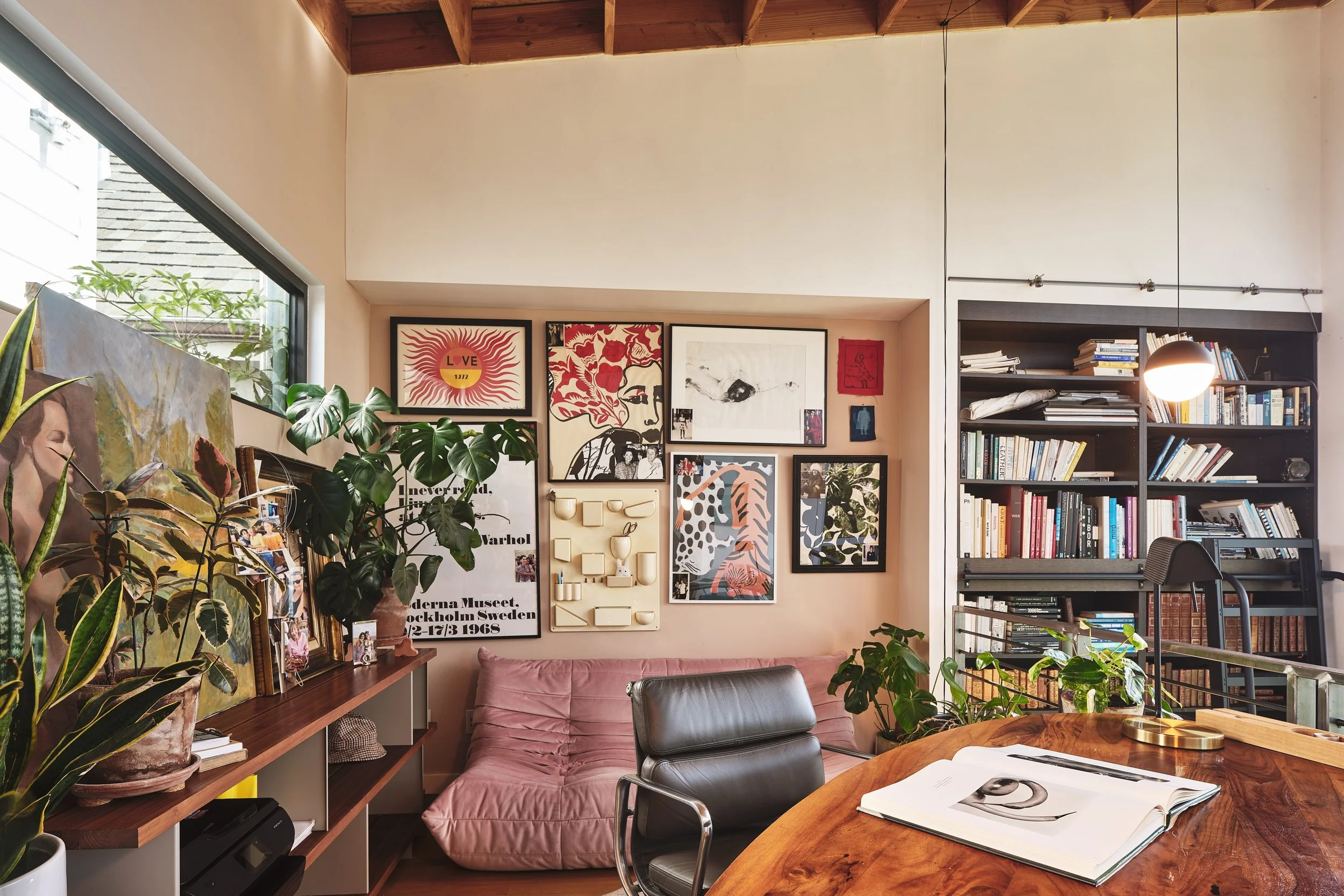 Interior of a stylish home office or living space with wooden ceiling, framed artwork on wall, bookshelf filled with books, plants, a table with open notebook, and a pink cushion on a seating area.