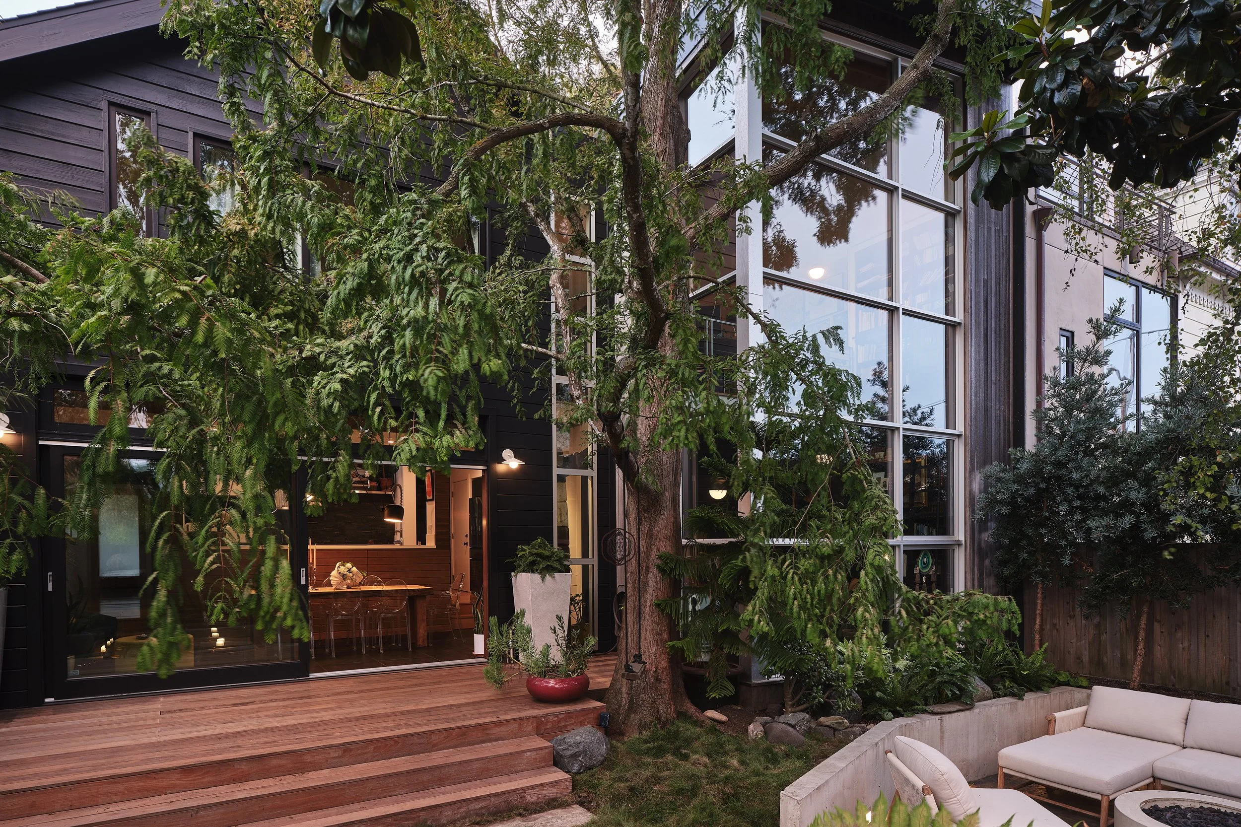 A modern house with large glass windows, a wooden deck, and lush green trees and plants in the backyard.