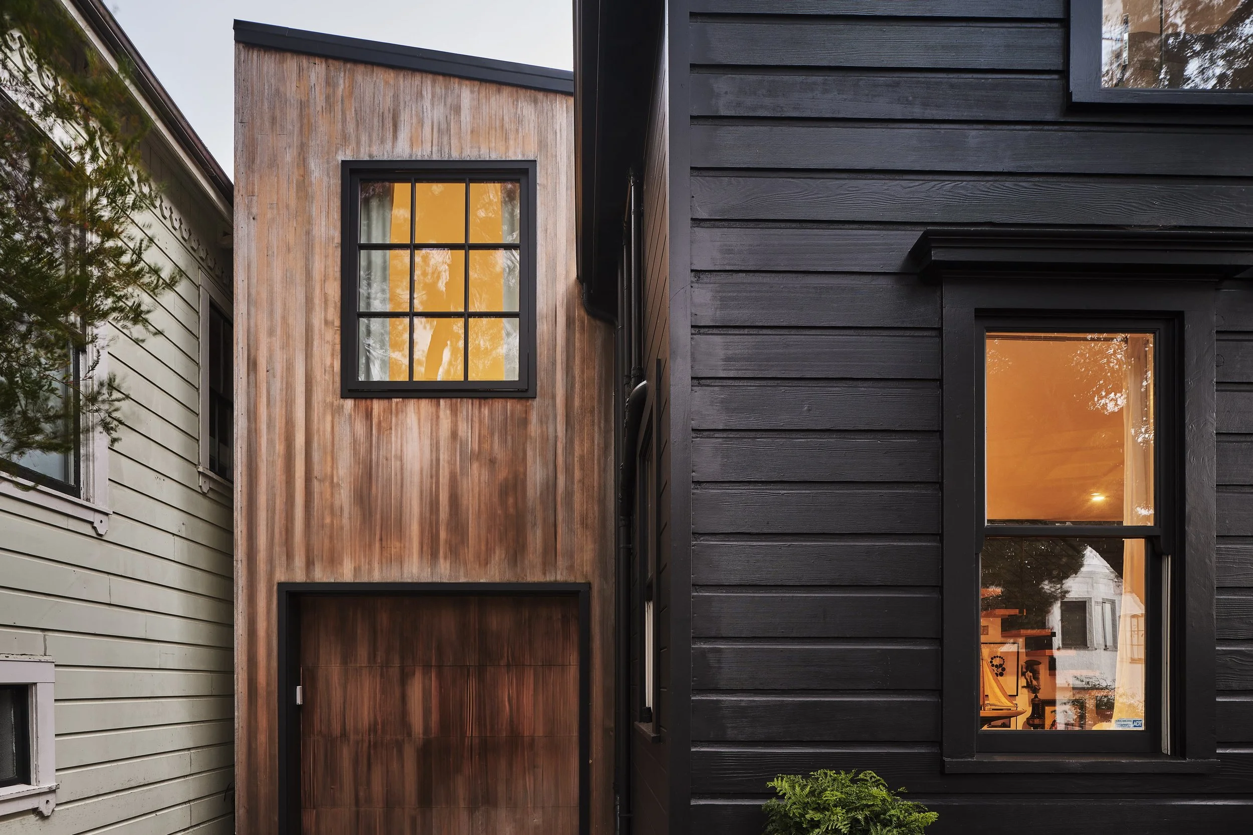 Close-up of a modern house with three different exterior wall styles: light green horizontal siding on the left, vertical wooden planks in the middle, and black horizontal wood siding on the right, each with windows reflecting trees and sky.