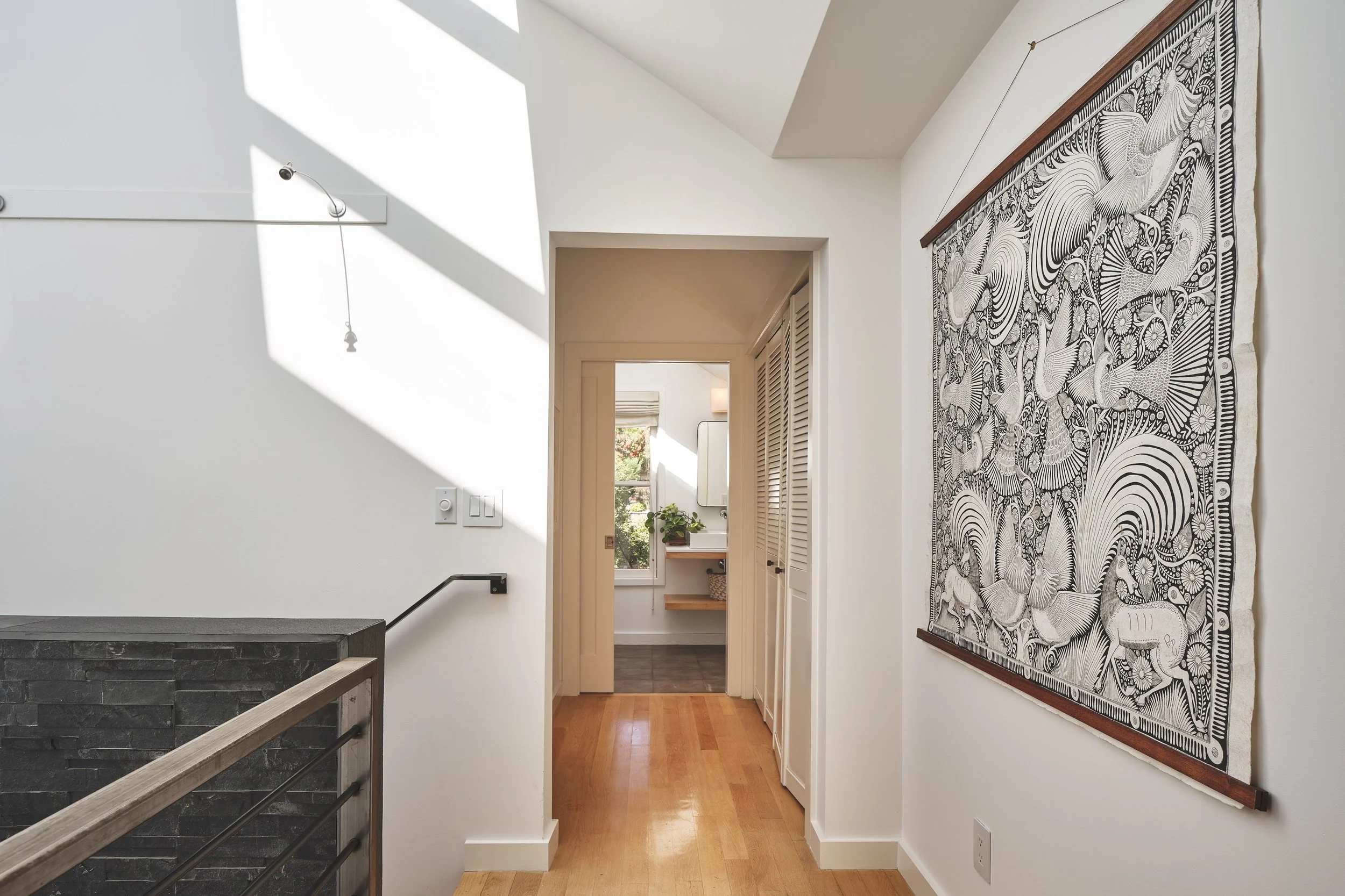 A hallway with wooden flooring, a large black-and-white artwork of birds on the right wall, and a staircase with a stone railing on the left. There are light switches and a small white cover on the wall, with sunlight coming through a window, illumin