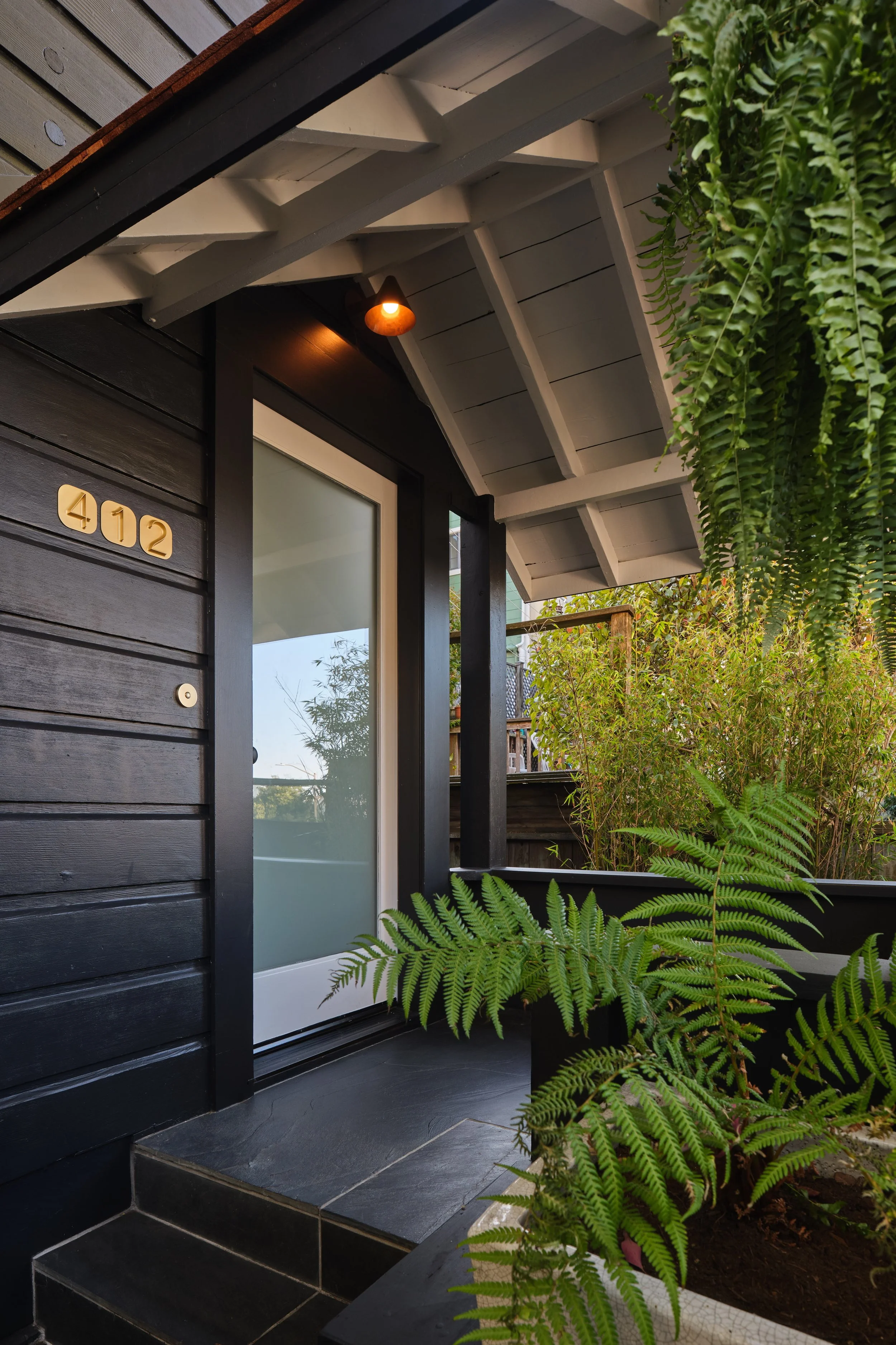 The front porch of a house with black siding, a frosted glass door, a small porch light, house number 402, and lush green plants including ferns.