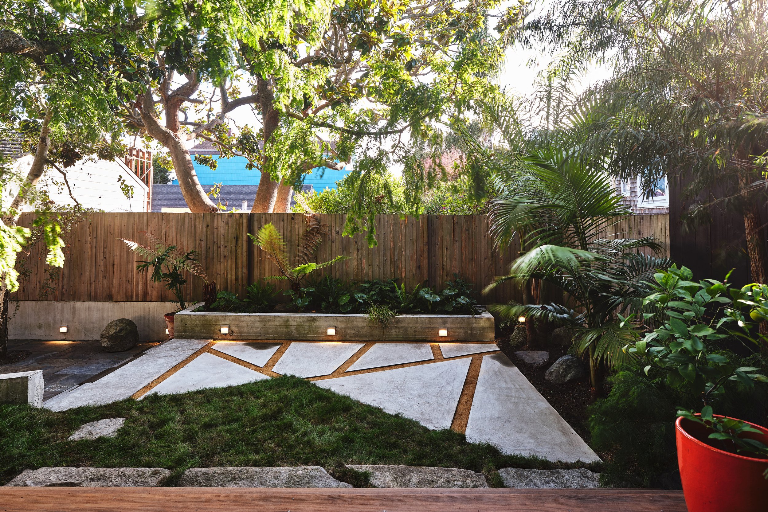 A backyard garden with a wooden fence, lush green trees and plants, a stone pathway, and outdoor lighting at dusk.