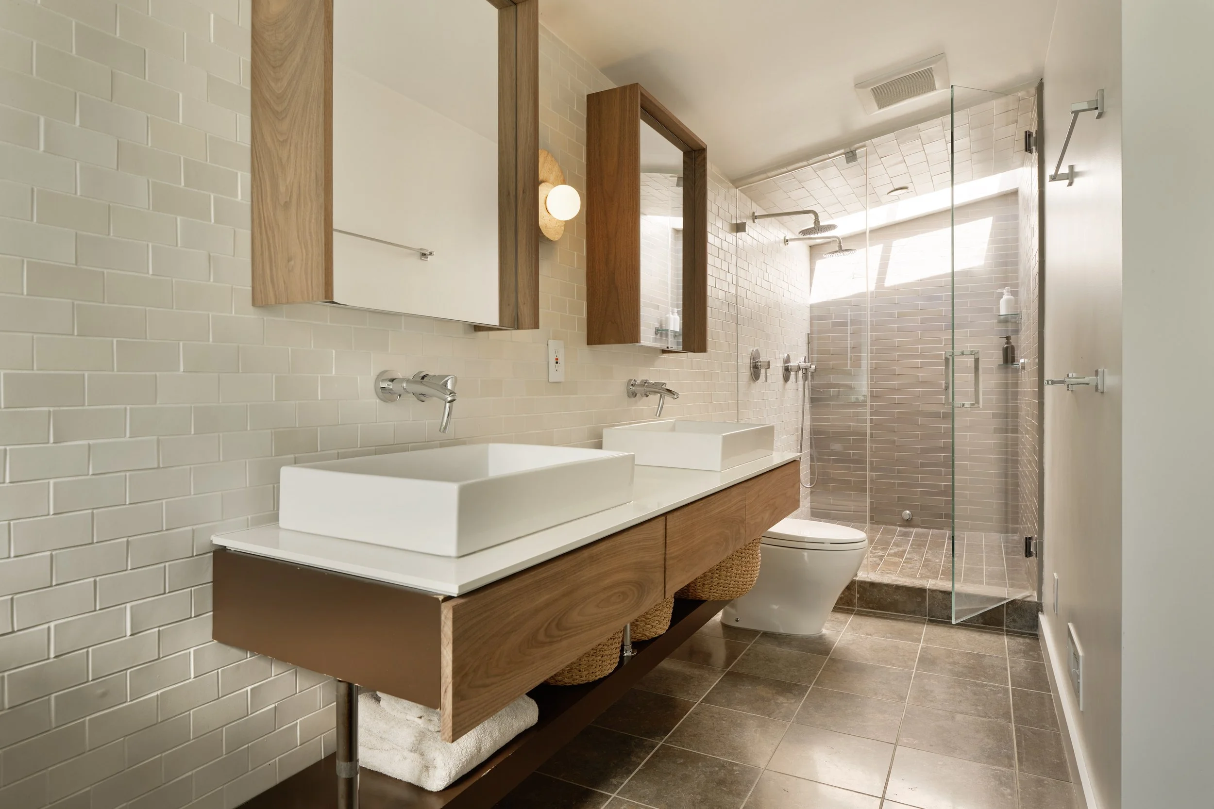 Modern bathroom with double sinks, large mirrors, wooden cabinetry, and a glass-enclosed shower with a rain showerhead, beige tile walls, and a dark tiled floor.