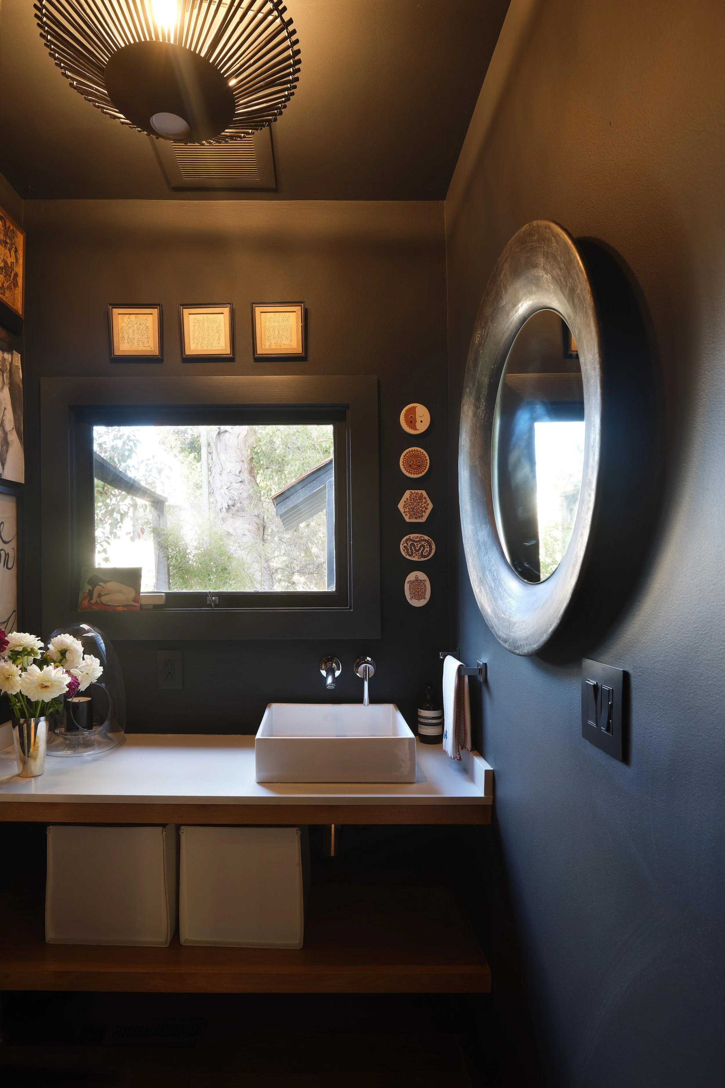 A modern bathroom with a white countertop, square sink, and a single faucet. Above the sink is a large round mirror with a metallic frame. To the left of the sink, there is a bouquet of white and purple flowers in a vase, and a framed picture. The window behind the sink shows a tree outside, with sunlight streaming in. The walls are dark gray, decorated with framed artwork and circular wall art. To the right of the mirror, a small towel hangs from a hook, and two black electrical switches are on the wall.