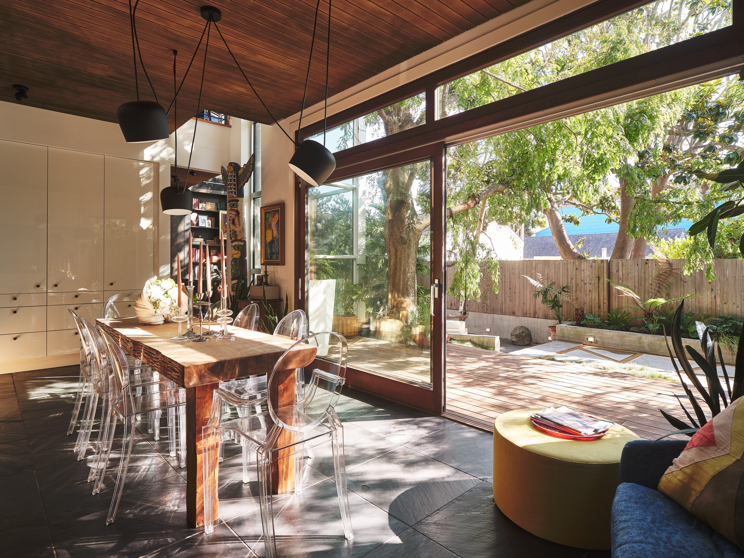 Modern dining area with a wooden table and transparent chairs, large windows showing a backyard with trees, and a wooden ceiling with hanging lamps.