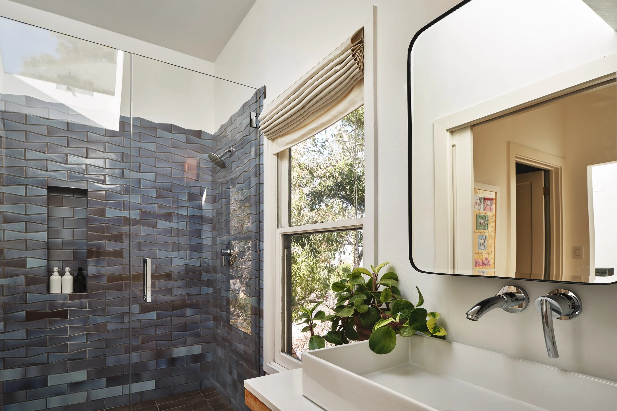 A modern bathroom with a walk-in shower featuring blue geometric tiles, a large mirror, a white sink with a silver faucet, a window with a beige Roman shade, and a potted plant.