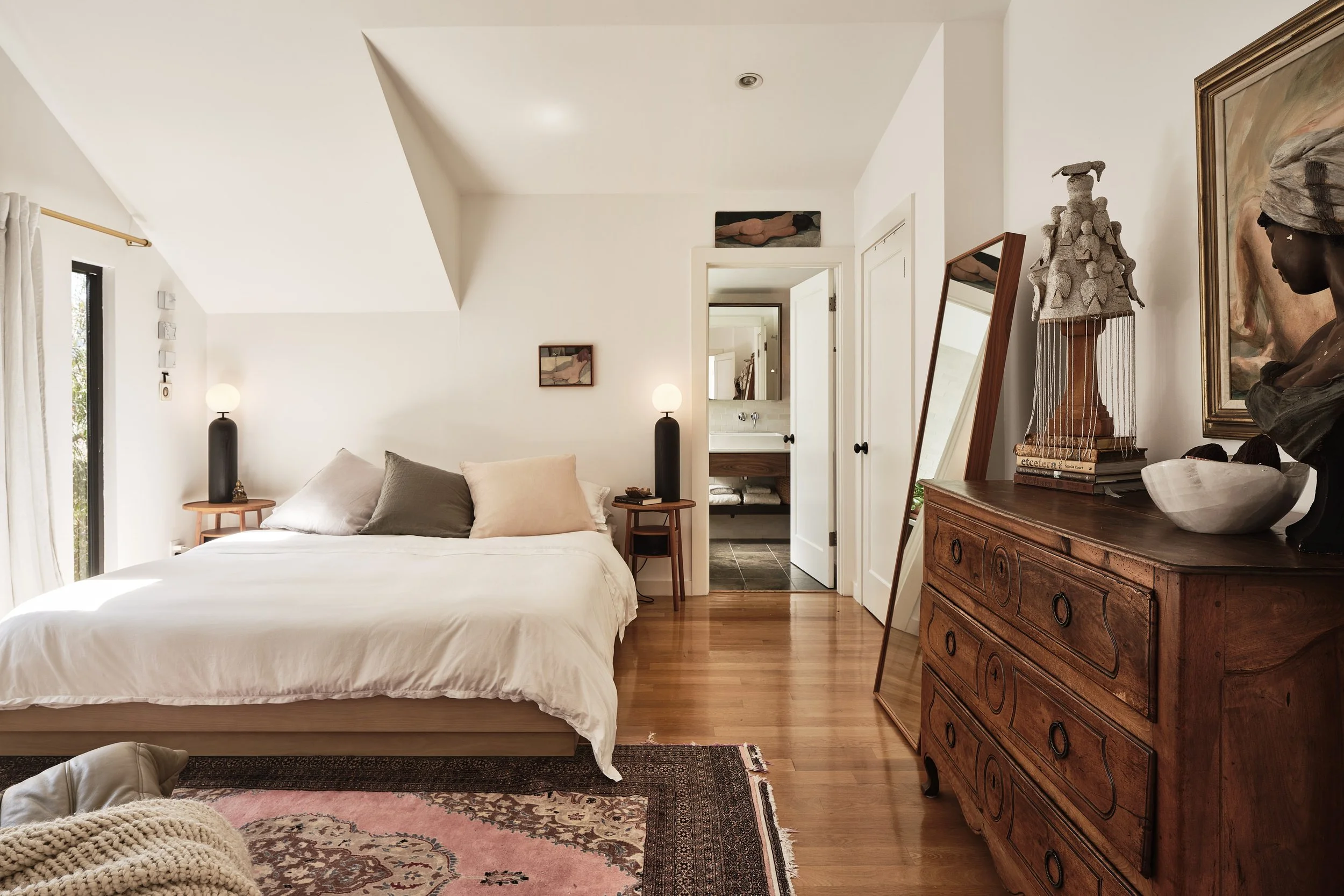 A bedroom with a bed, two nightstands with lamps, an open bathroom door, and a wooden dresser with various decorative items, including sculptures, books, and a framed painting on the wall.