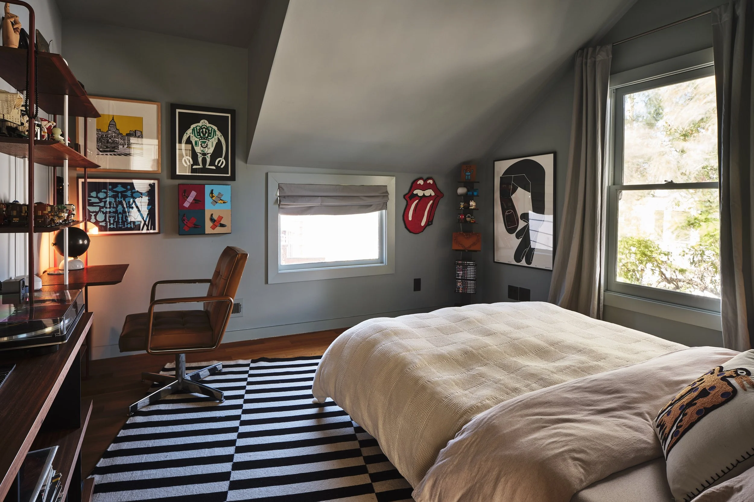 A bedroom with a bed, a window with curtains, and framed artwork on the walls. There is a patterned black and white rug on the wooden floor, and a tan swivel chair in the corner.