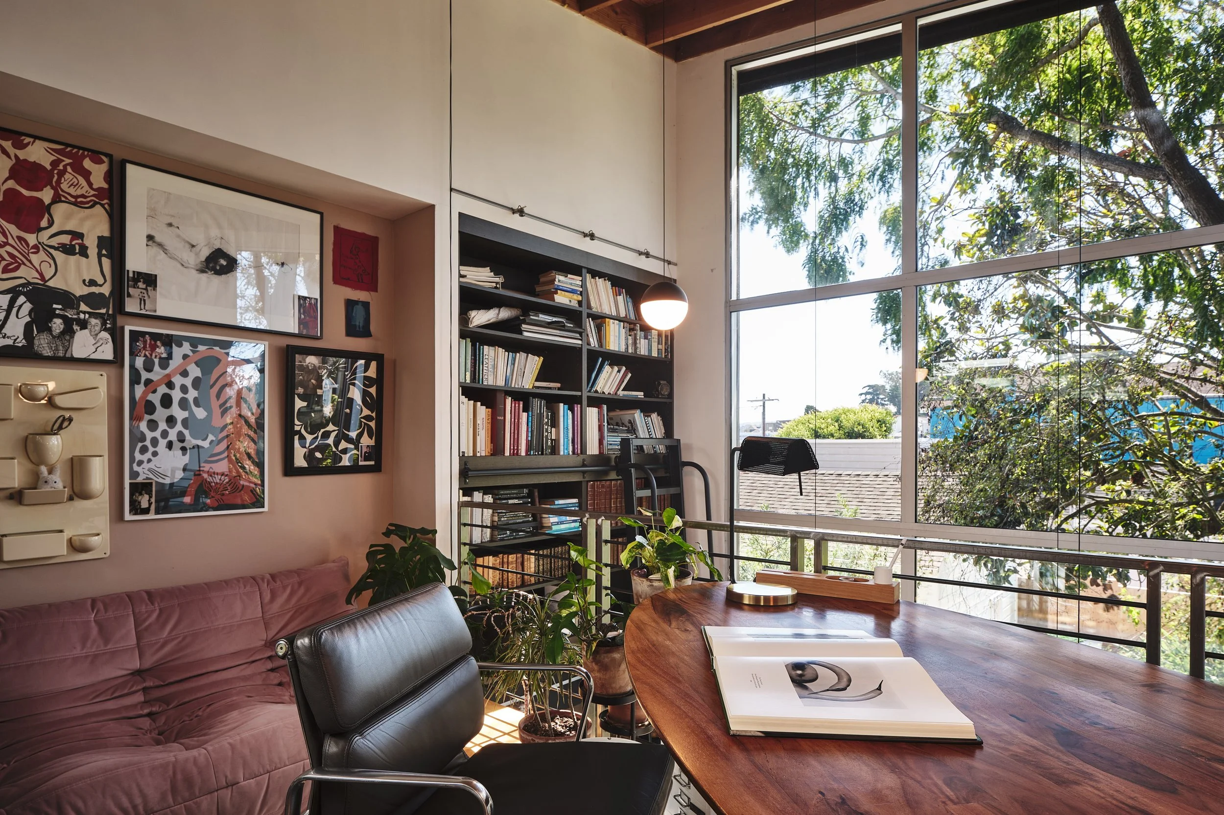A cozy home office with a wooden desk, bookshelves filled with books, framed artwork on the wall, a large window showing trees outside, and a pink velvet sofa.