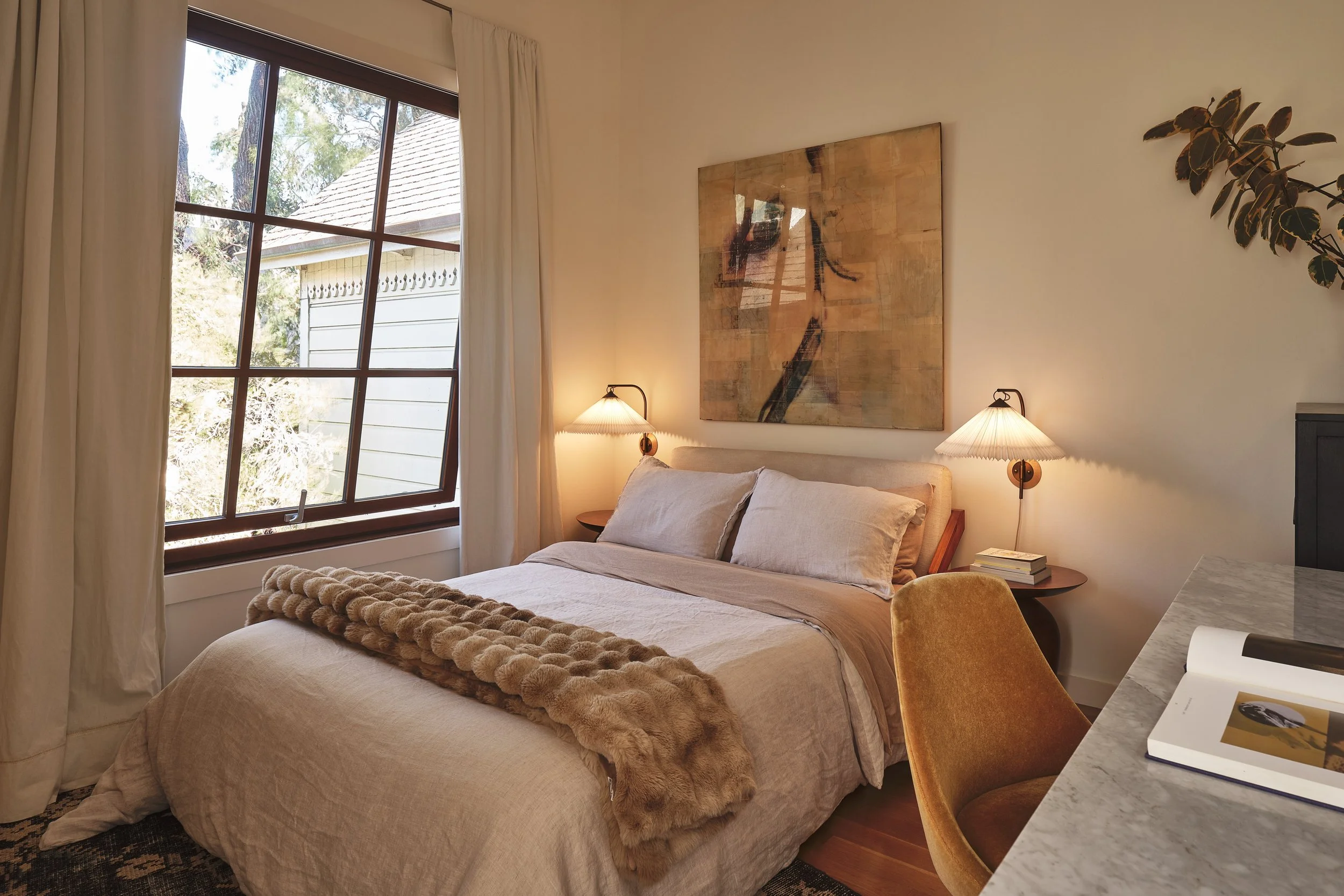 A cozy bedroom with a bed, beige bedding, and a brown fur blanket. Two wall-mounted bedside lamps with beige shades are above the bed. A large abstract painting hangs on the wall, and a window with cream curtains lets in natural light, showing trees 