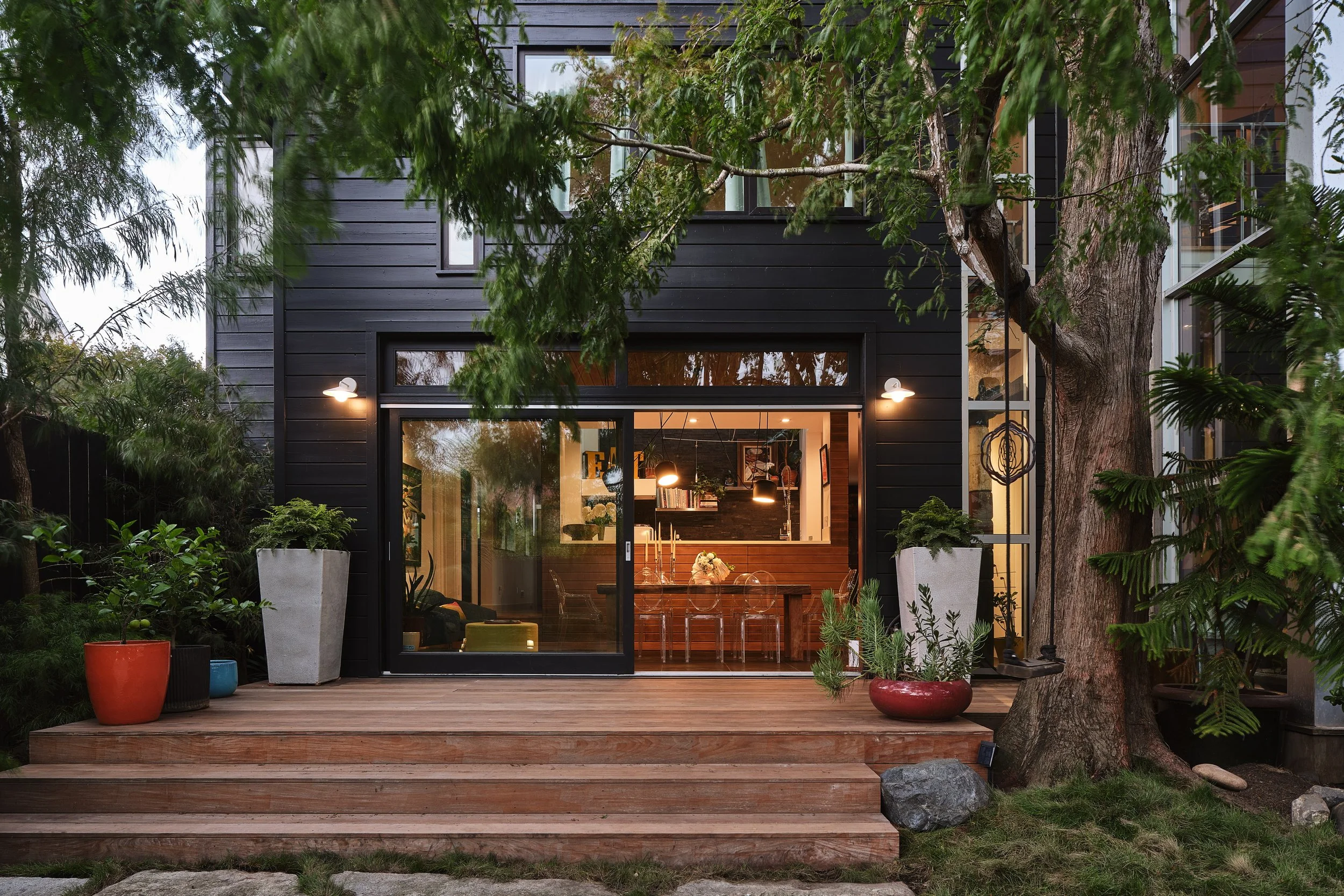 A modern black house has a large window and sliding glass door on a wooden deck, surrounded by green trees and potted plants outside, with interior lighting visible.