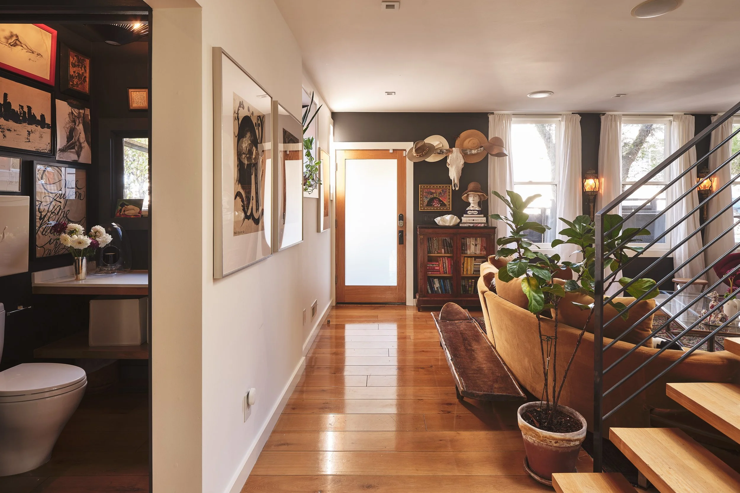 A cozy living room with wooden floors, a yellow couch, a tall green potted plant, and large windows with white curtains. The walls are decorated with various hats, artwork, and a cow skull. There is a small wooden bookshelf near the door, and a stair