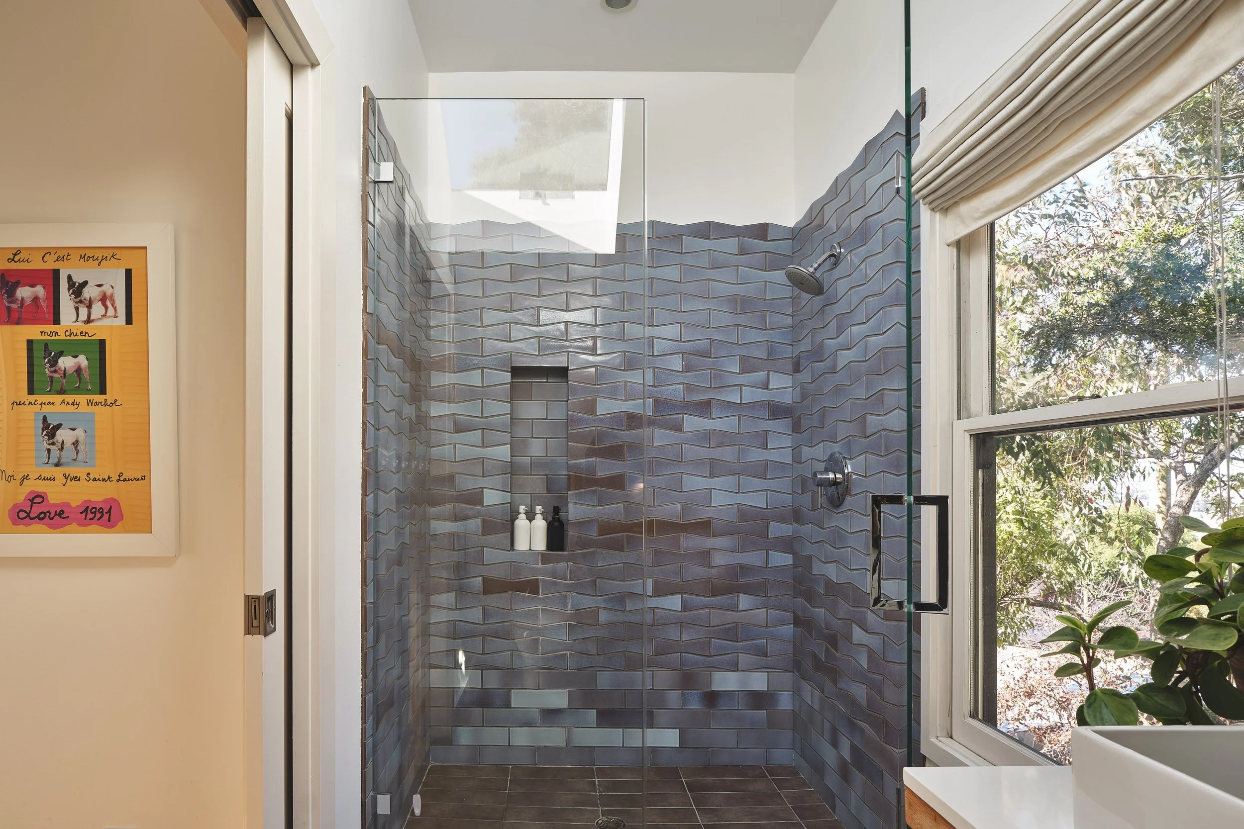 Modern shower with blue geometric tiles, clear glass door, and a skylight overhead. A built-in niche holds shampoo and soap bottles. Large window with a view of trees and a partially open white window shade, with a small potted plant nearby.