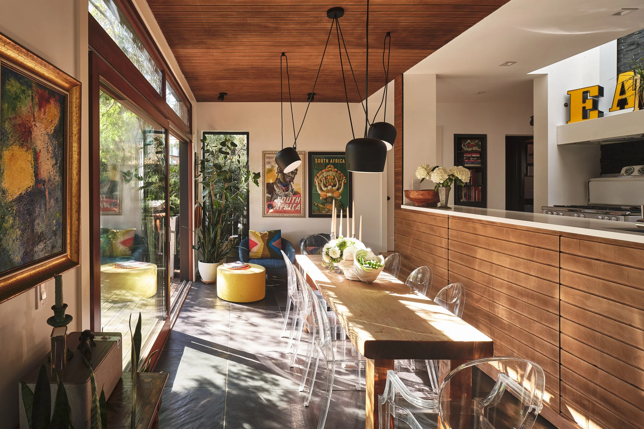 A bright, modern dining area with a wooden ceiling and large glass doors leading to an outdoor space. The room is decorated with artwork, plants, and a table set with flowers and candles. There are transparent chairs around the wooden dining table an