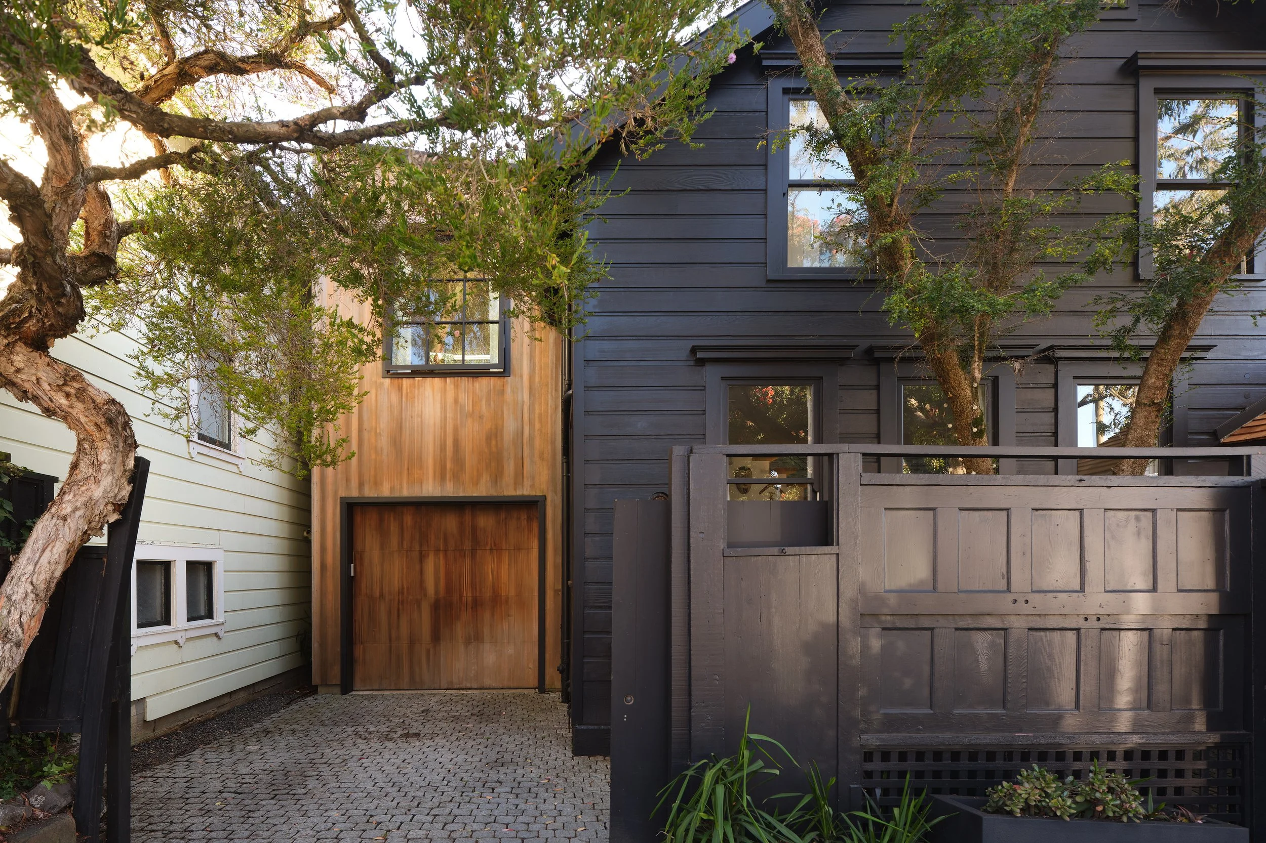 A modern black wooden house with large windows partially obscured by tree branches and leaves, neighboring to a white house with smaller windows, and a cobblestone pathway in front.