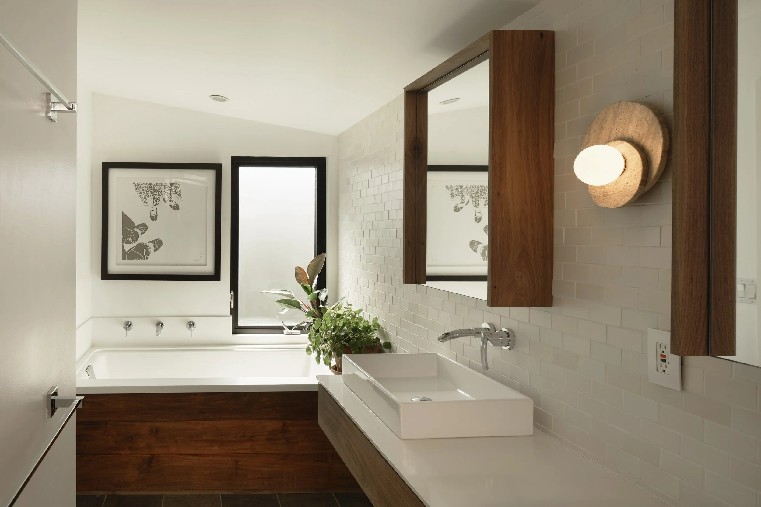 Modern bathroom with white brick walls, a wooden frame mirror, a white sink with a chrome faucet, a light fixture, framed artwork, and a window with frosted glass.