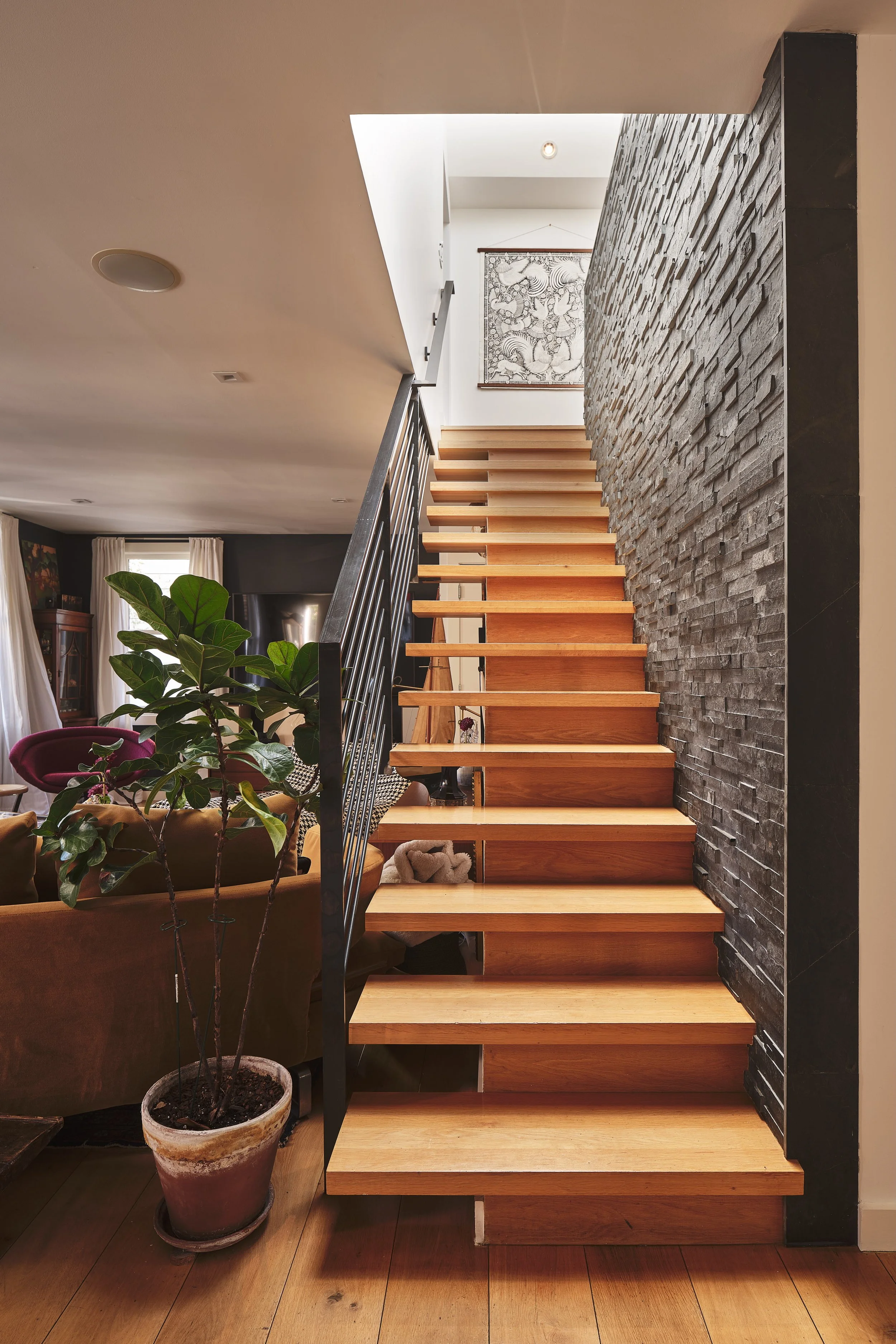 Wooden staircase with black metal railing, adjacent to a textured black stone wall, in a modern living space.