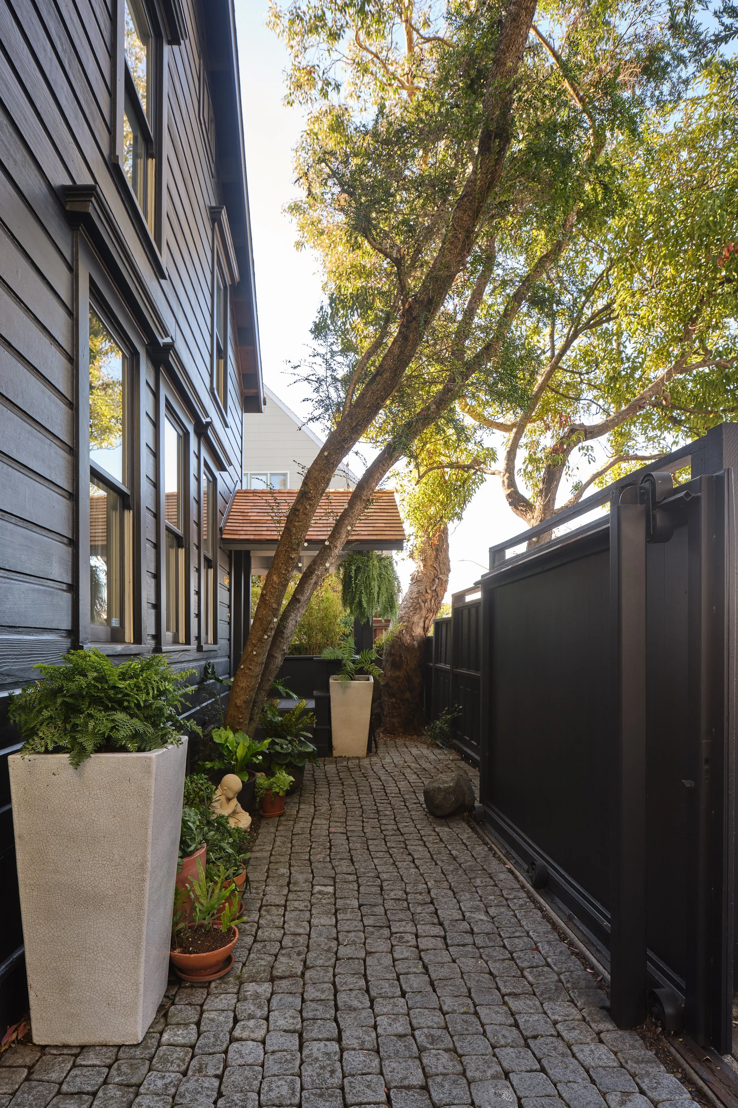 View of a narrow cobblestone alleyway between a black wooden house and a black fence, with various potted plants and a small statue, and a large tree with green leaves overhead.