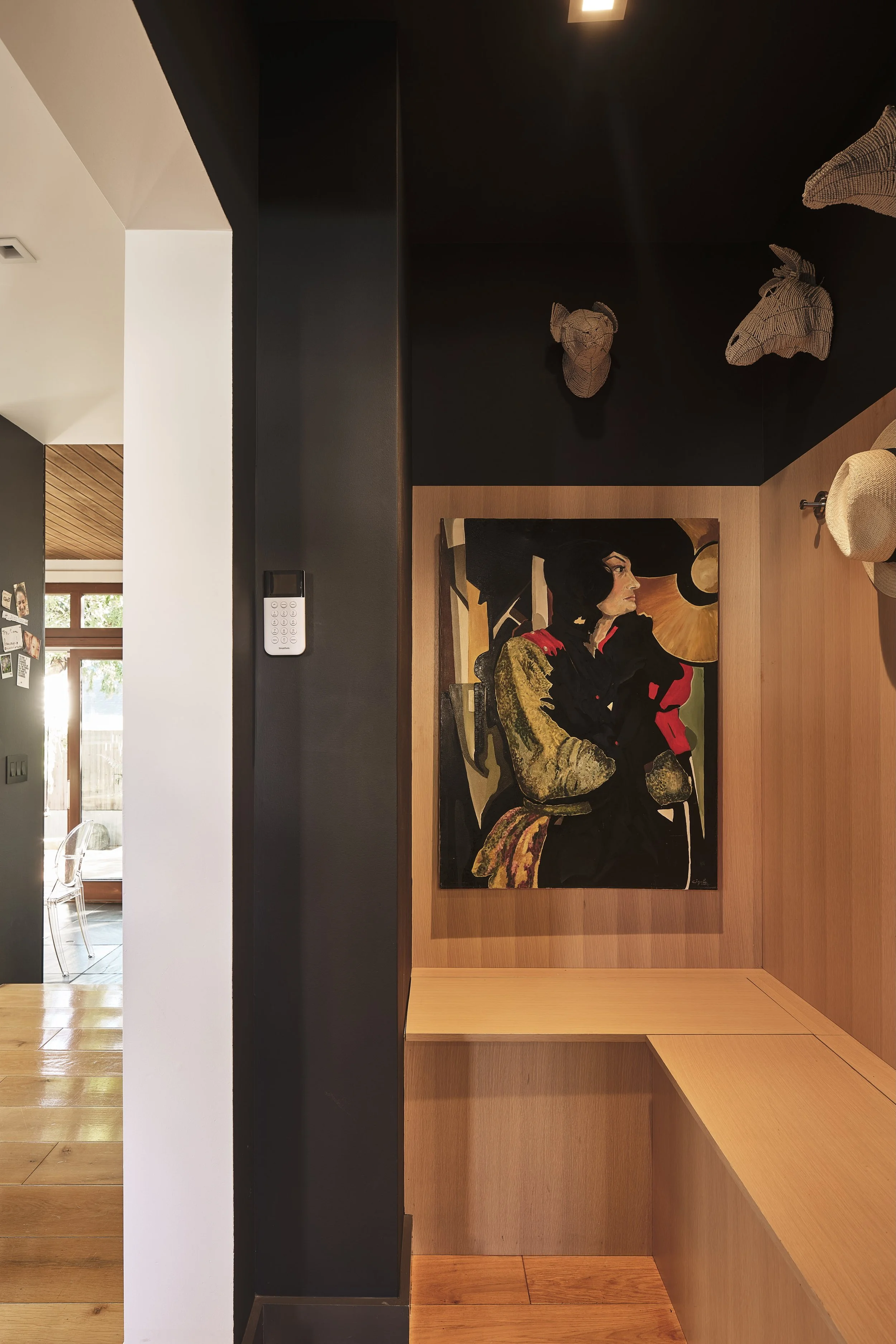 Interior of a modern room with a black wall, a framed painting of a woman with black hair and a red bow, three wireframe animal head sculptures on the black wall, a beige hat on a shelf, and a keypad on the black wall. Part of the adjacent room with 